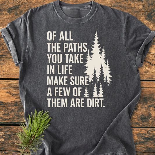 Of All The Paths (John Muir) - Vintage Wash Tee features a charcoal-gray design with an inspiring nature-themed slogan and stylized pine trees, laid on a wooden surface with fresh pine needles.