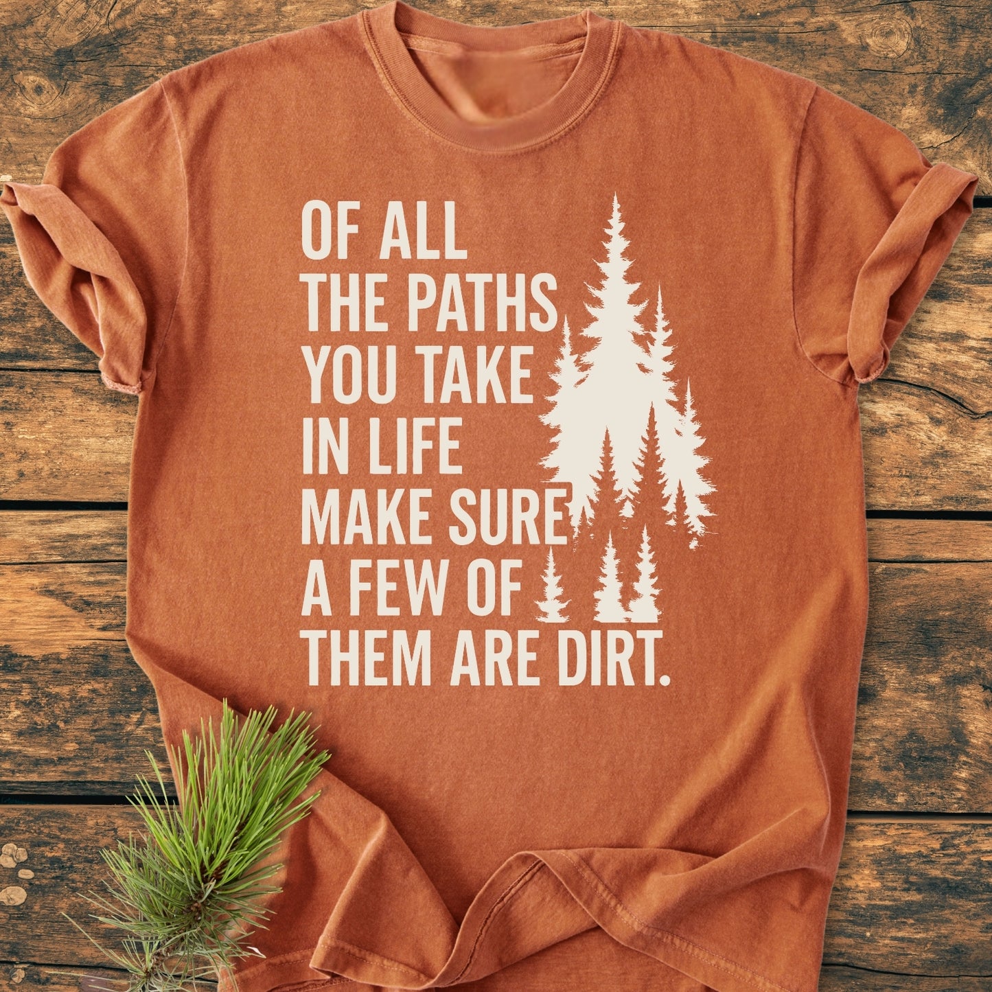 Of All The Paths (John Muir) - Vintage Wash Tee, rust-orange with bold white lettering and pine trees design, laid flat on wood. Cozy, relaxed fit, 100% cotton, garment-dyed for comfort.