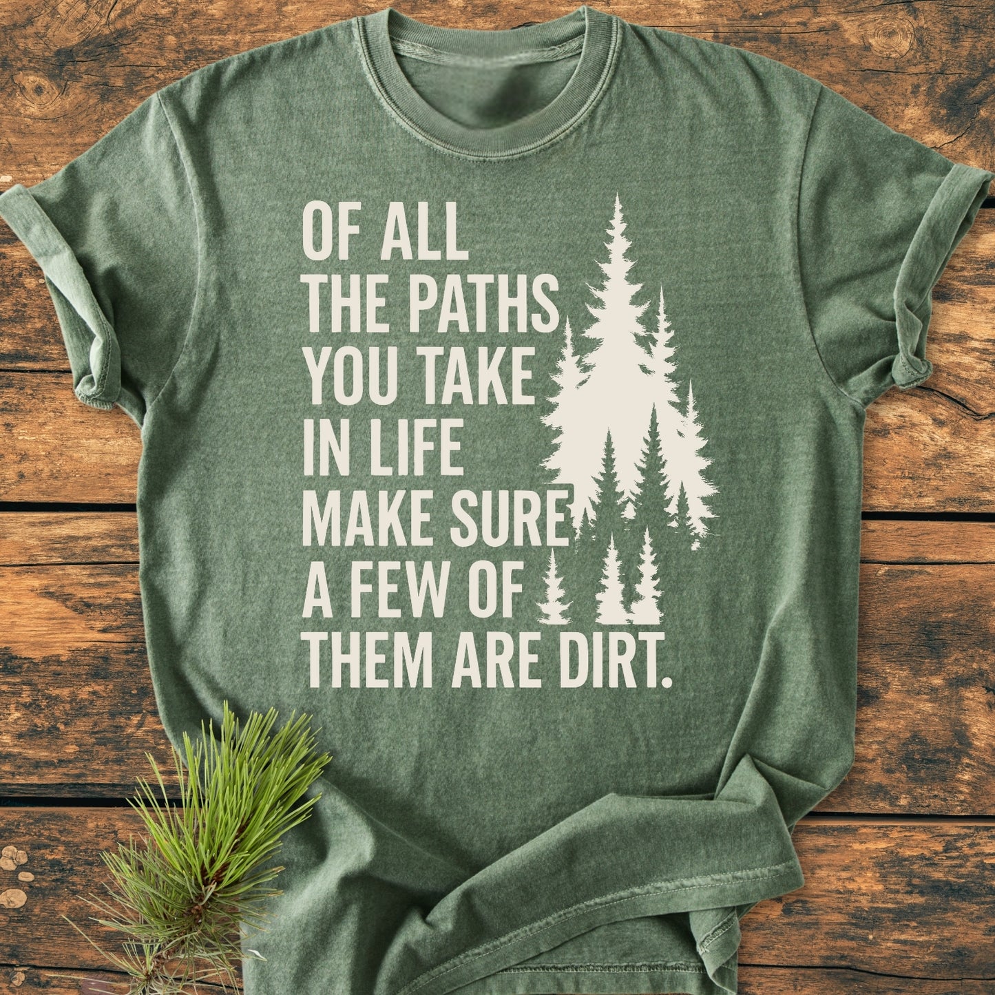 The Of All The Paths (John Muir) - Vintage Wash Tee features a white pine tree graphic and slogan on an olive-green heathered T-shirt, laid on wood, with a pine branch beside it.