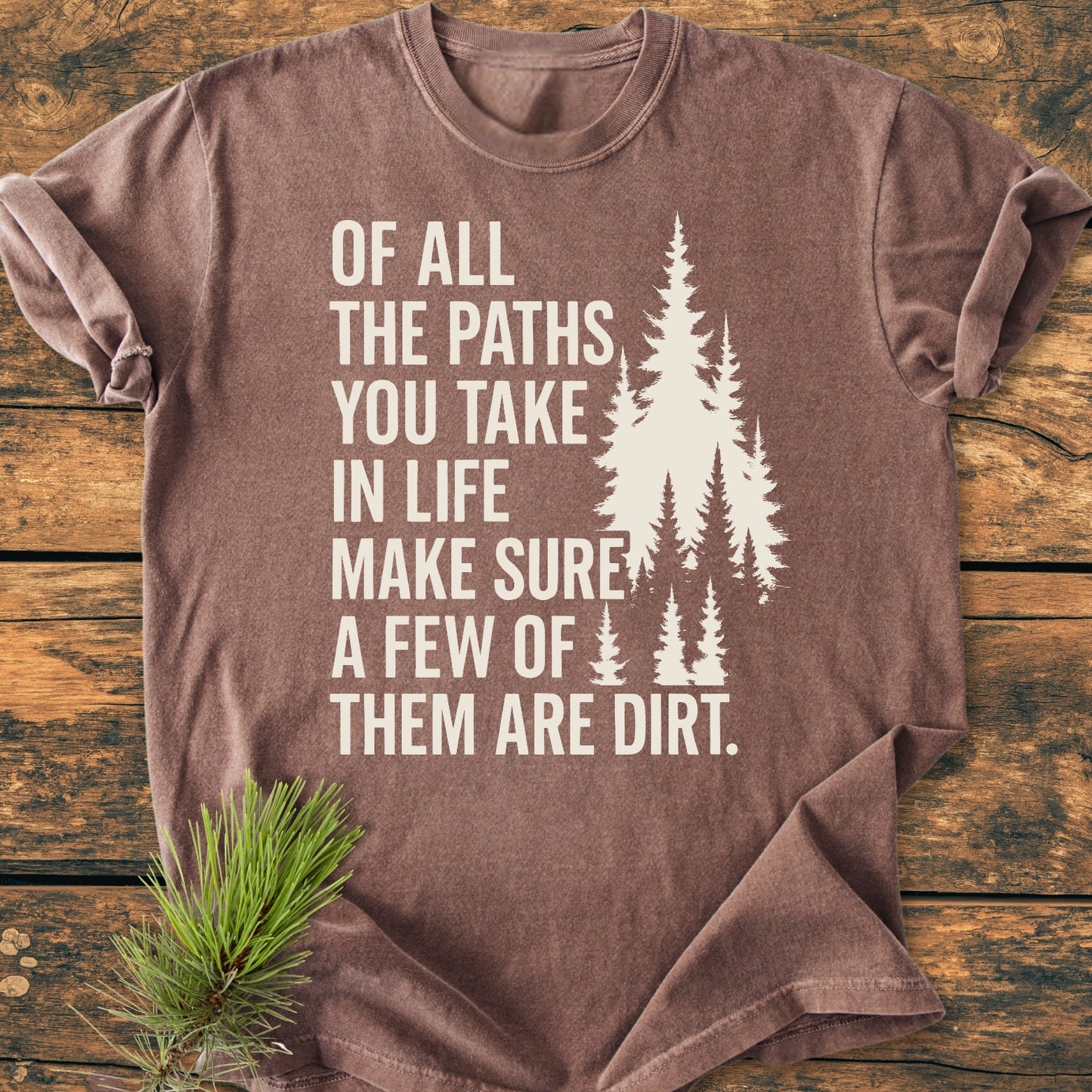 Of All The Paths (John Muir) - Vintage Wash Tee featuring pine tree silhouettes and slogan, laid on wood with pine needles. Made from soft, durable 100% ring-spun cotton, offering a relaxed fit.
