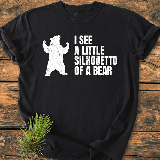 The I See A Little Silhouetto Of A Bear - Vintage Wash Tee features a distressed bear graphic with playful text on a black T-shirt, styled with a pine branch on a wooden surface.