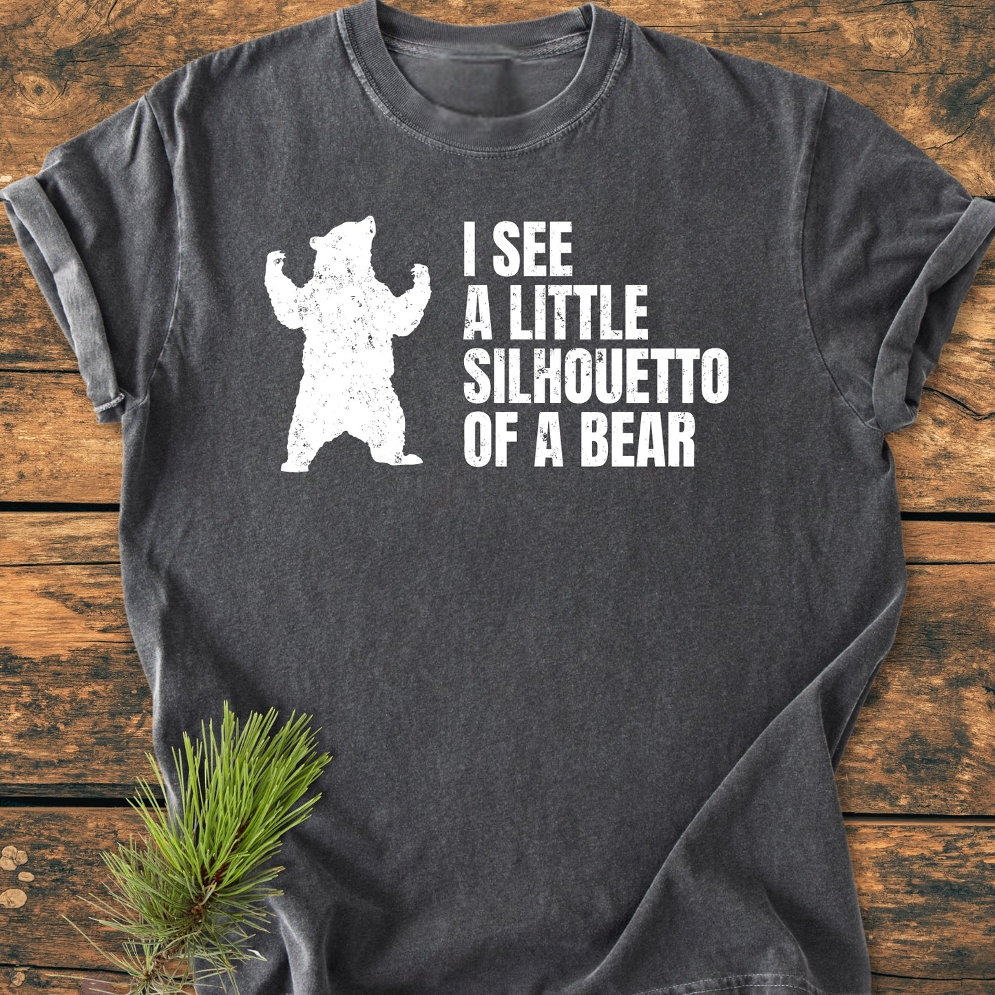 Charcoal-gray I See A Little Silhouetto Of A Bear - Vintage Wash Tee featuring a bear silhouette and playful text in distressed white ink, laid flat on a wooden surface with an evergreen sprig nearby.