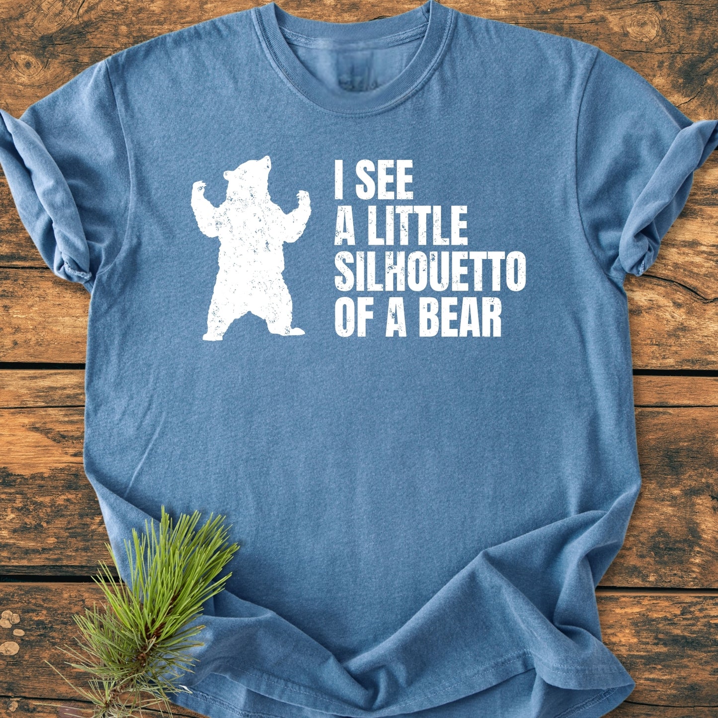 Heather-blue I See A Little Silhouetto Of A Bear - Vintage Wash Tee with distressed white bear silhouette and playful text, laid flat on rustic wood, accented by a pine sprig.