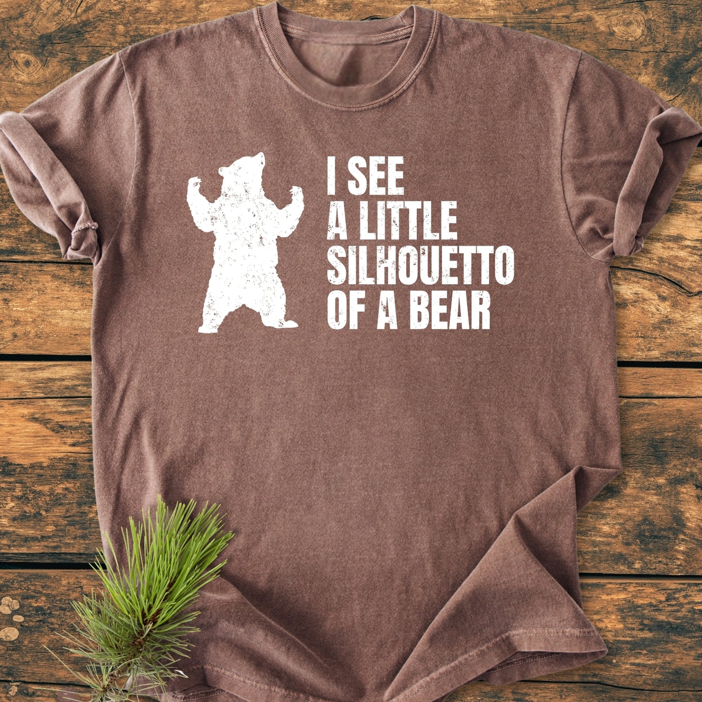 I See A Little Silhouetto Of A Bear - Vintage Wash Tee features a distressed bear silhouette and playful text on a brown T-shirt, laid on wood with a pine sprig accent.