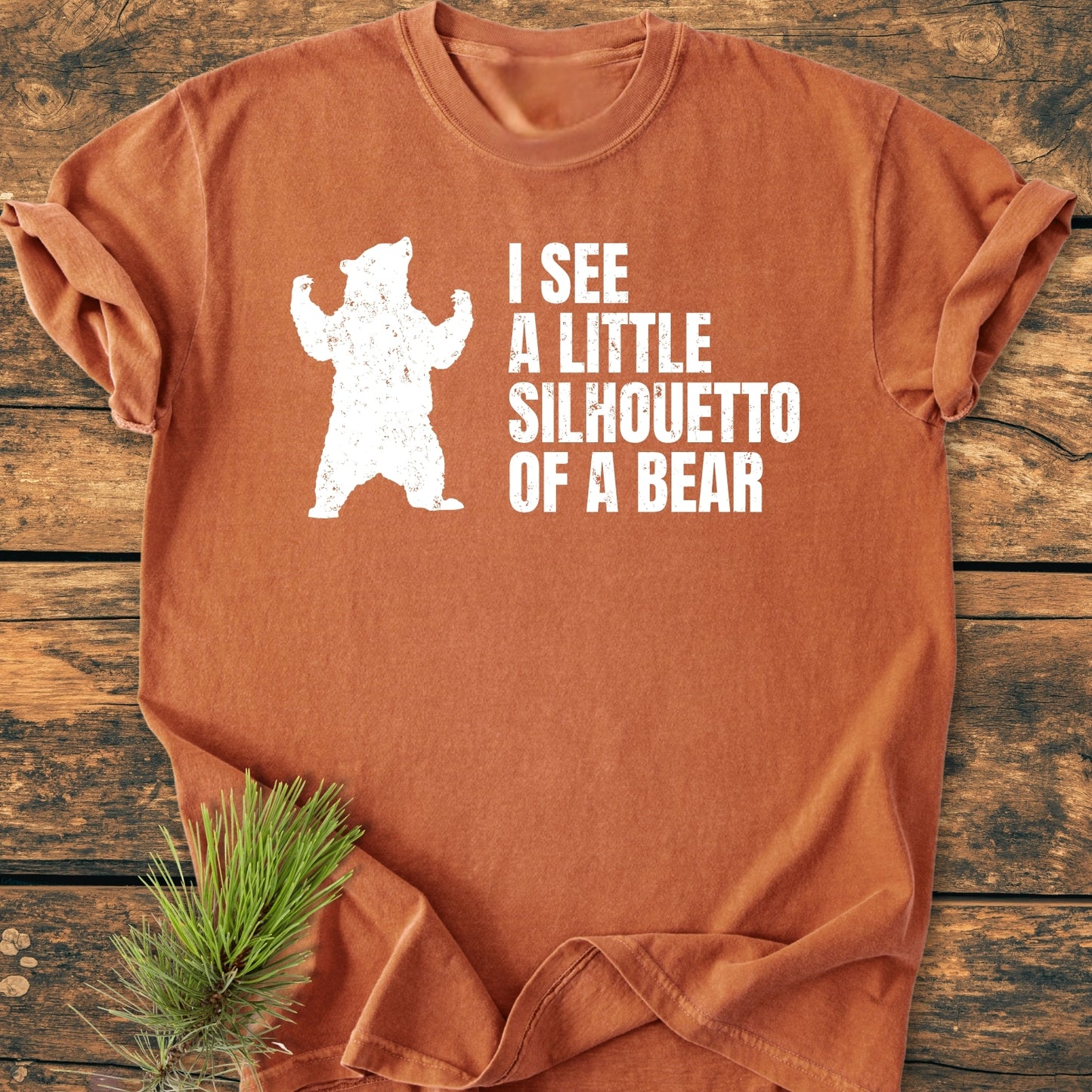 I See A Little Silhouetto Of A Bear - Vintage Wash Tee: Rust-orange T-shirt with a distressed white bear graphic and playful text, laid flat on wood with a pine sprig for styling.