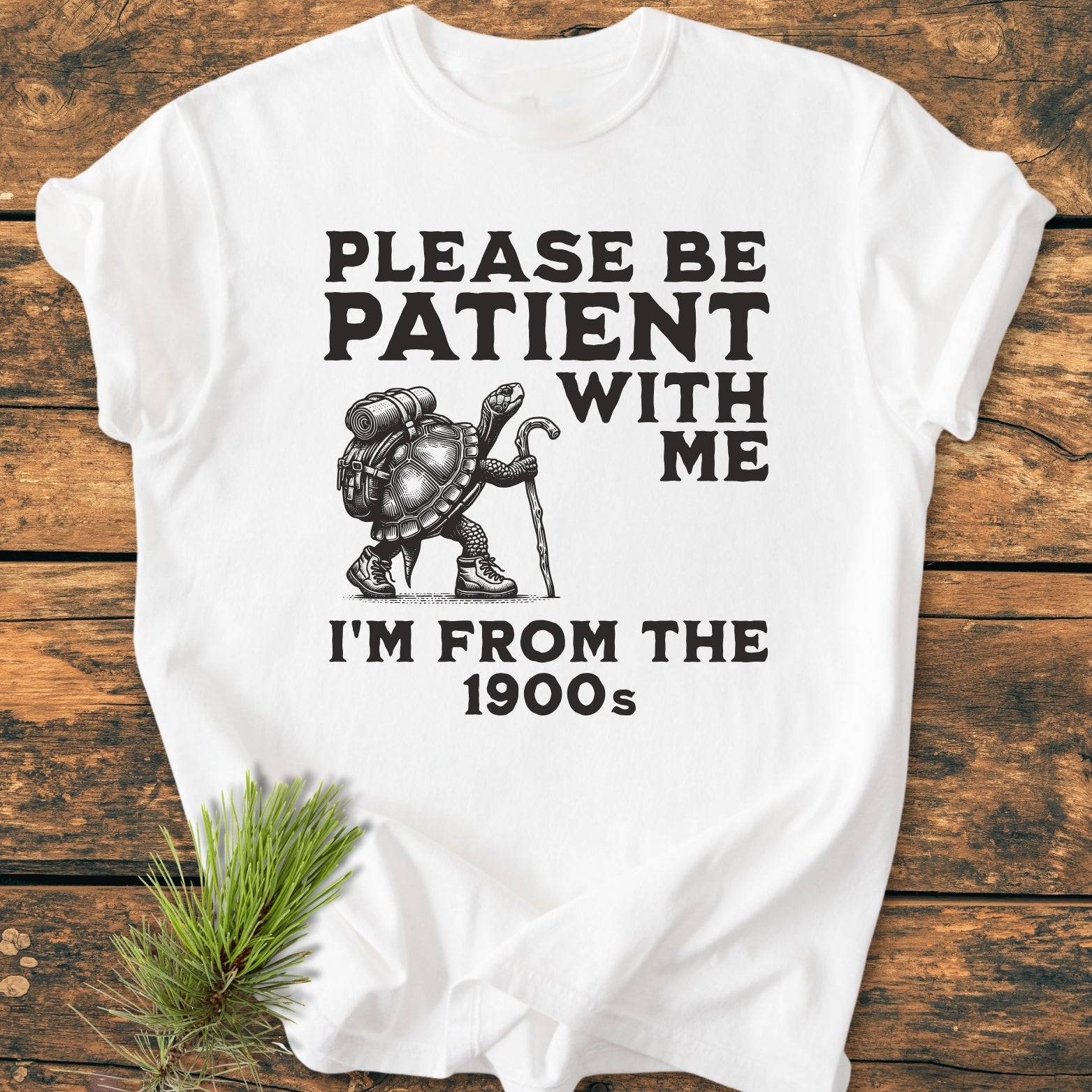 Please Be Patient I'm From The 1900s - Vintage Wash Tee features a turtle in hiking gear with humorous text on a soft, durable white T-shirt, laid on a wooden surface.