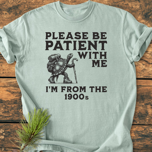 Please Be Patient I'm From The 1900s - Vintage Wash Tee features a tortoise in hiking gear with bold text, laid on a wooden surface, emphasizing its relaxed fit and soft, durable cotton fabric.