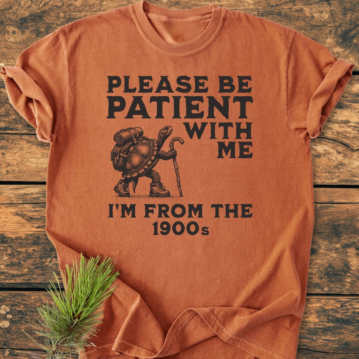 Please Be Patient I'm From The 1900s - Vintage Wash Tee features a turtle illustration with hiking gear on a burnt-orange background, laid on wood, highlighting its quirky, nostalgic appeal.