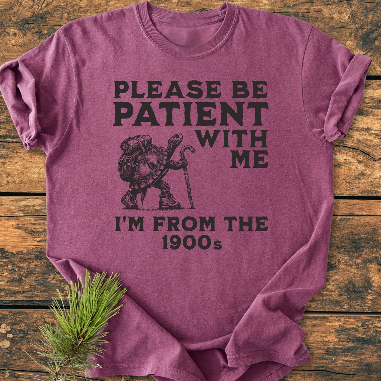Please Be Patient I'm From The 1900s - Vintage Wash Tee featuring a black turtle graphic with hiking gear and slogan, folded neatly on a wooden surface, showcasing its relaxed fit and durable stitching.