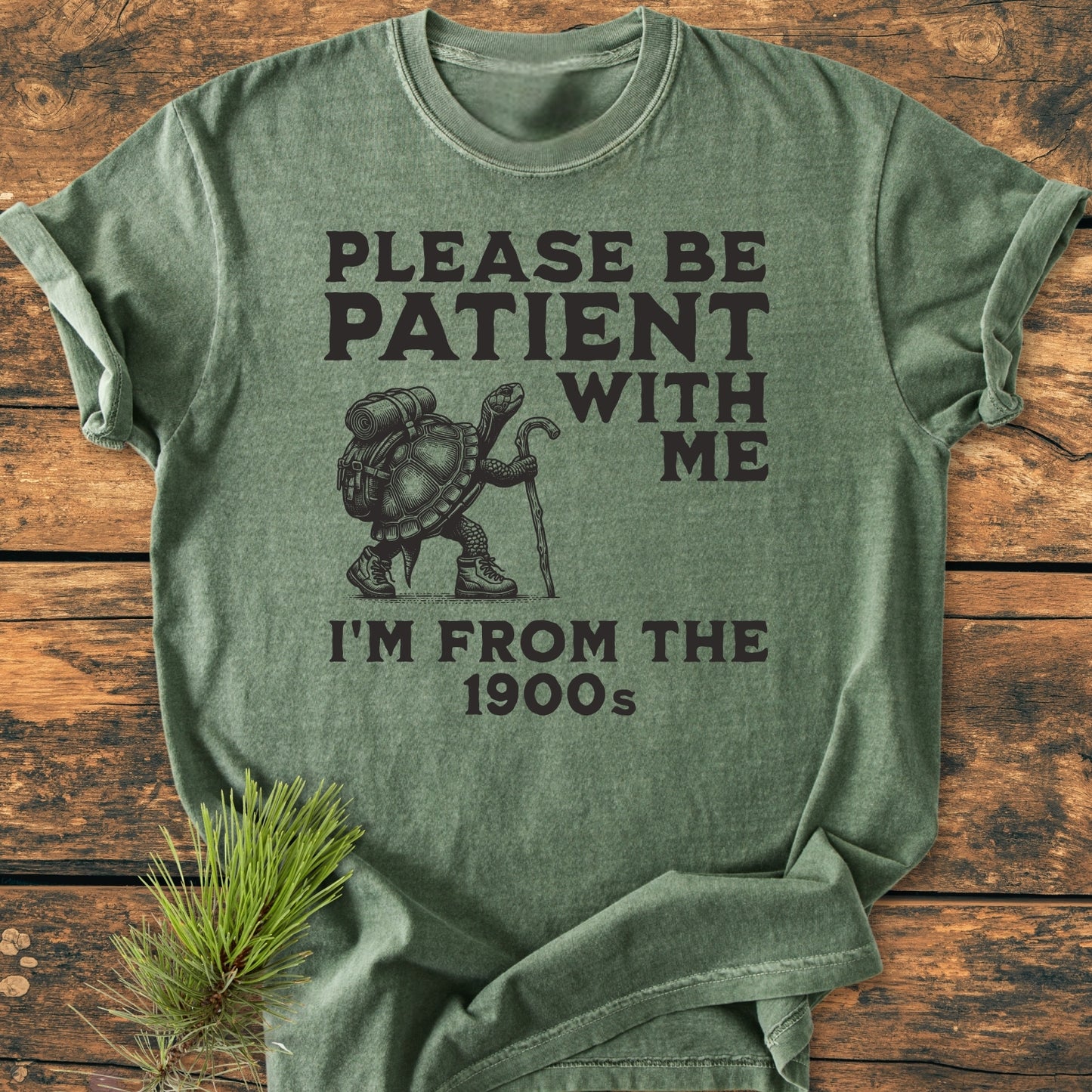 Green heather T-shirt featuring a cartoon turtle in hiking gear with the slogan “PLEASE BE PATIENT WITH ME, I’M FROM THE 1900s.” Relaxed fit, 100% ring-spun cotton, durable stitching.