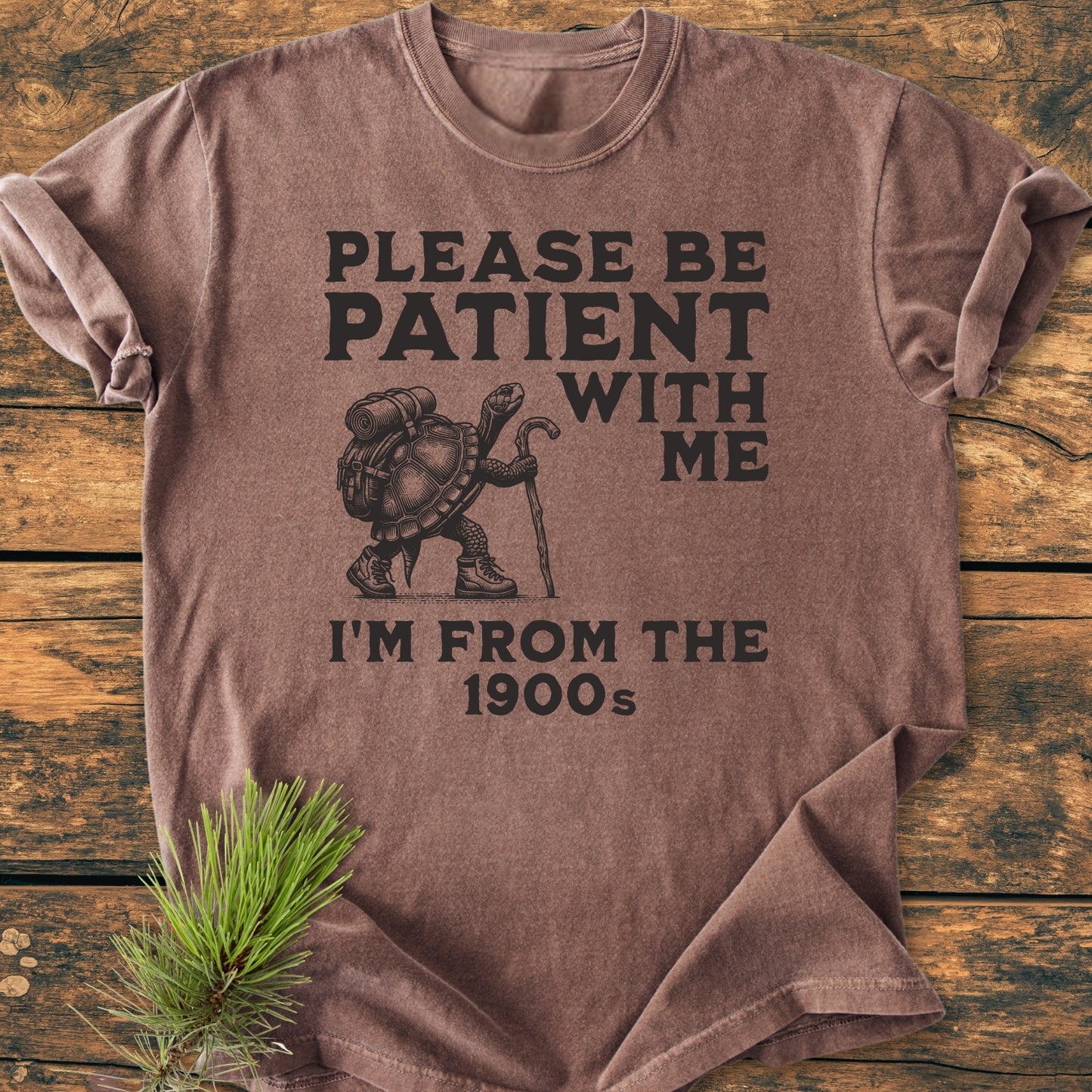 Please Be Patient I'm From The 1900s - Vintage Wash Tee featuring a hiking tortoise with boots and a backpack, laid on a wooden surface with a pine sprig.
