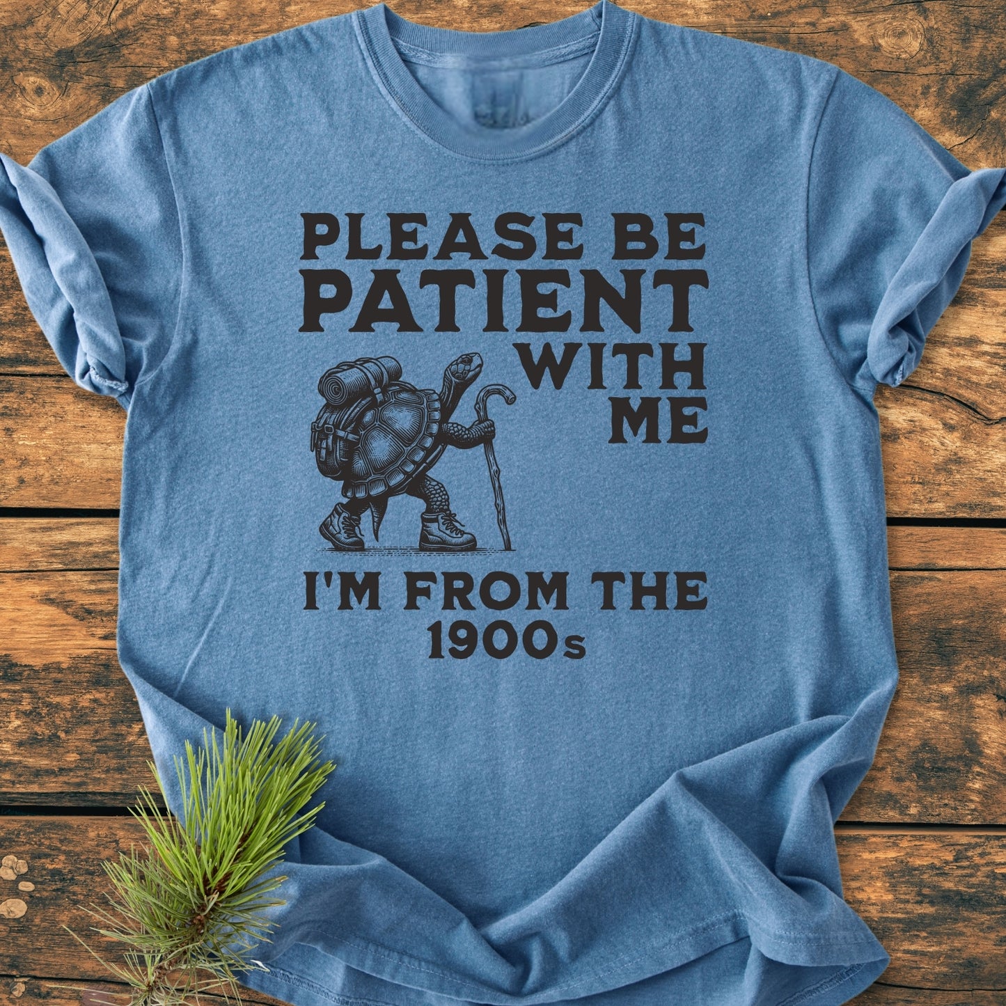 Heather-blue Please Be Patient I'm From The 1900s - Vintage Wash Tee featuring a hiking turtle graphic, laid flat on wood with a pine sprig. Bold lettering and relaxed fit for comfort.
