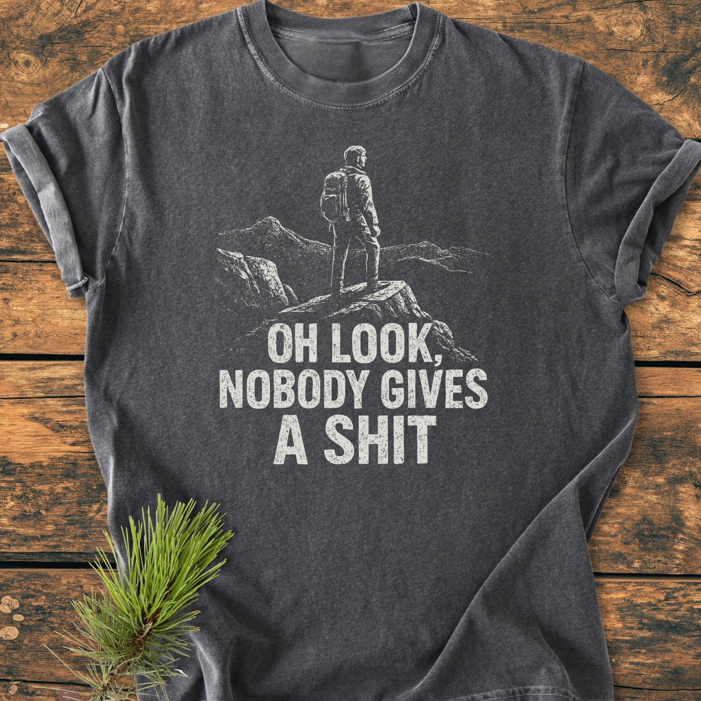 Oh Look - Vintage Wash Tee featuring a hiker silhouette and cheeky text in white on charcoal-gray fabric, displayed with rolled sleeves on wood, complemented by a pine sprig nearby.