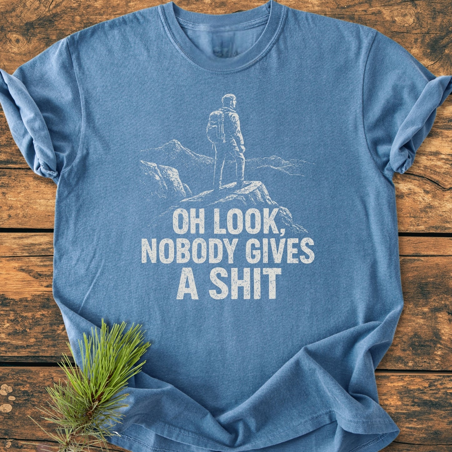 Heather-blue Oh Look - Vintage Wash Tee featuring a white sketch of a person on a rocky outcrop, with bold text. Laid on wood, highlighting the T-shirt's relaxed fit and soft-washed texture.