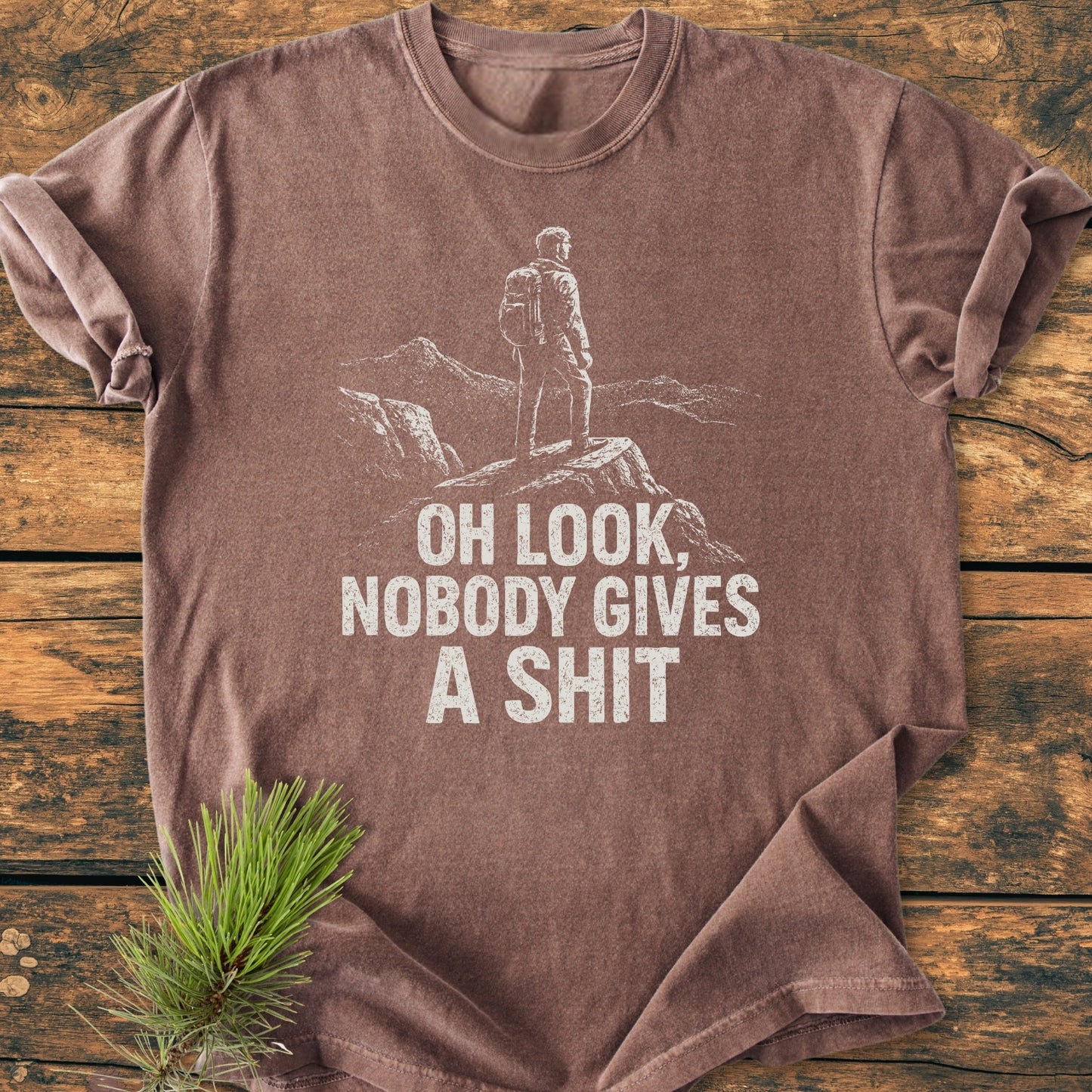 Oh Look - Vintage Wash Tee: Brown short-sleeved T-shirt with white line-art of a backpacker on rocks, bold distressed text, styled with pine sprig on wooden surface.