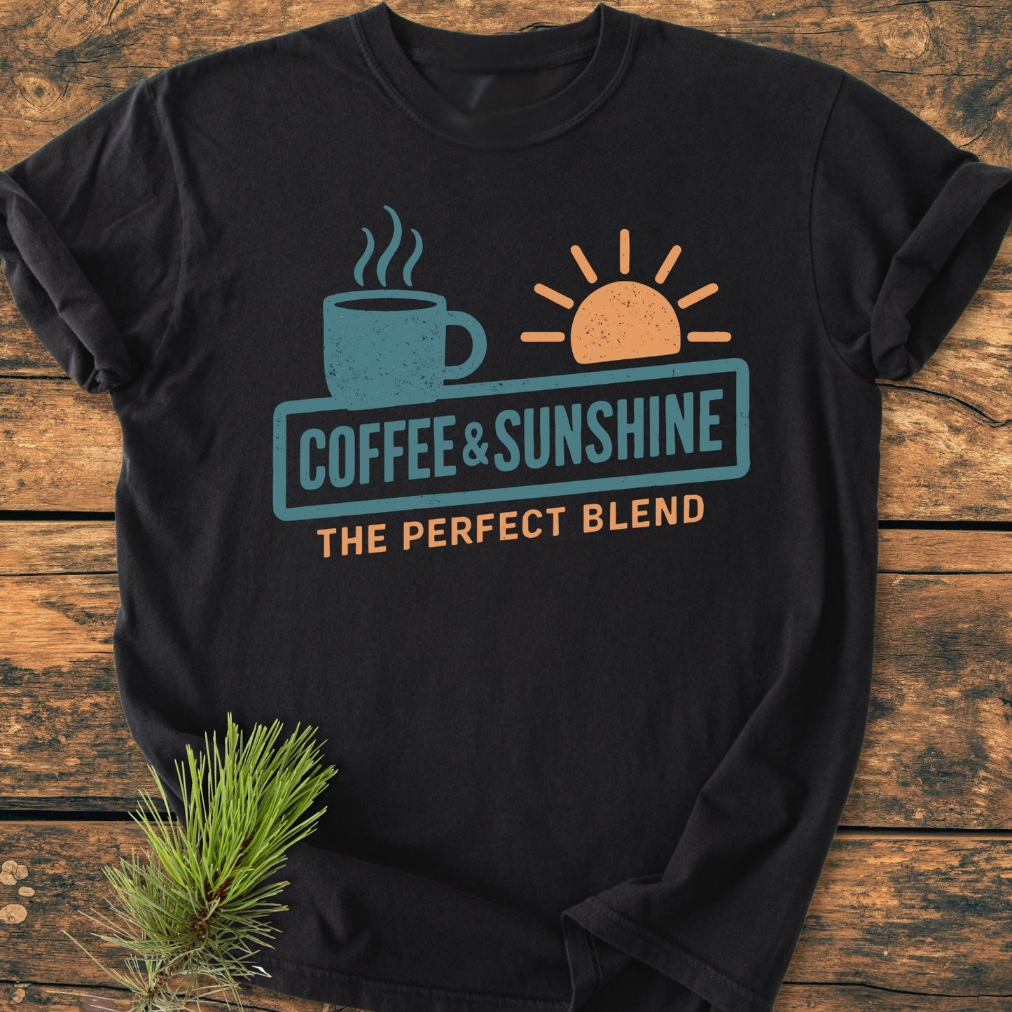 Coffee And Sunshine, Perfect Blend - Vintage Wash Tee: Black T-shirt with coffee mug and half-sun graphic, slogan beneath. Laid flat on wood, pine sprig tucked in corner, showcasing relaxed fit and garment-dyed fabric.