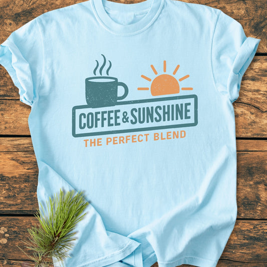 Light-blue Coffee And Sunshine, Perfect Blend - Vintage Wash Tee featuring a coffee mug and sun graphic, slogan, and pine sprig on a wooden surface. Made from soft, durable, pre-shrunk cotton.
