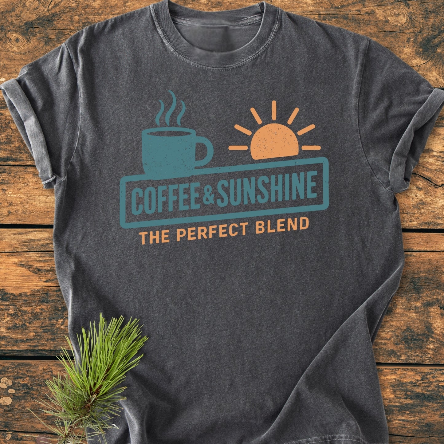 Coffee And Sunshine, Perfect Blend - Vintage Wash Tee features a charcoal-gray design with teal coffee mug, orange sun graphics, and text on soft-washed cotton, laid on rustic wood.