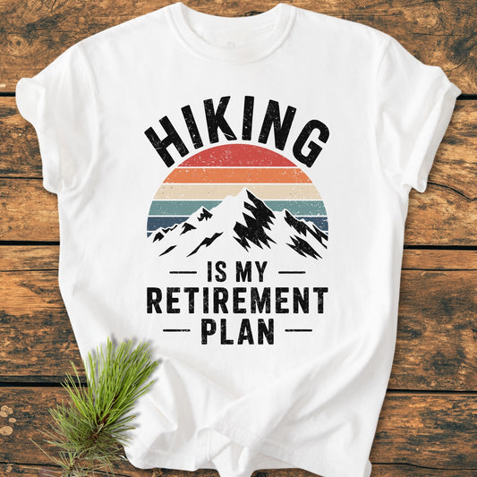 Hiking Is My Retirement Plan - Vintage Wash Tee featuring a retro mountain graphic and slogan. Laid on wood with pine sprig, showcasing its relaxed fit, soft-washed cotton fabric, and durable stitching.