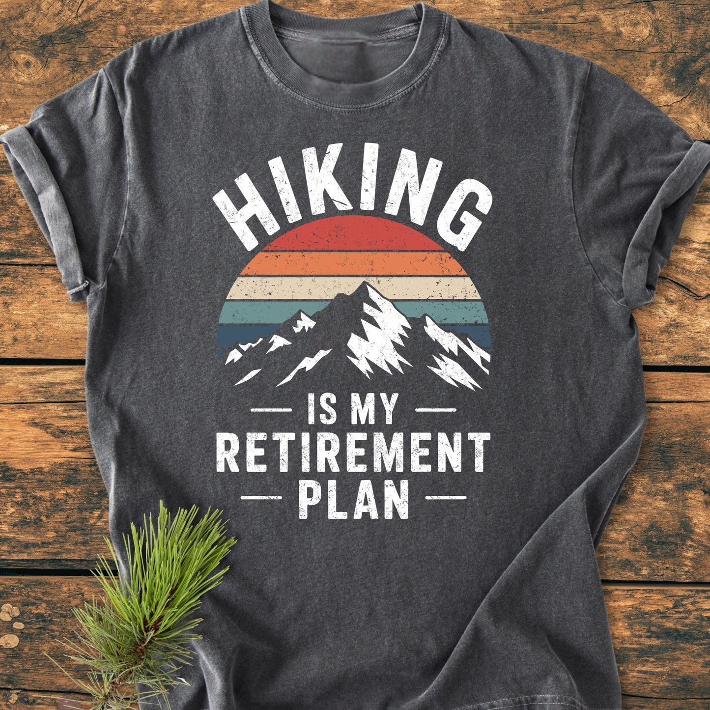 Charcoal-gray Hiking Is My Retirement Plan - Vintage Wash Tee features a mountain silhouette with a striped sun graphic and slogan, displayed on a wooden surface with a pine sprig beside it.