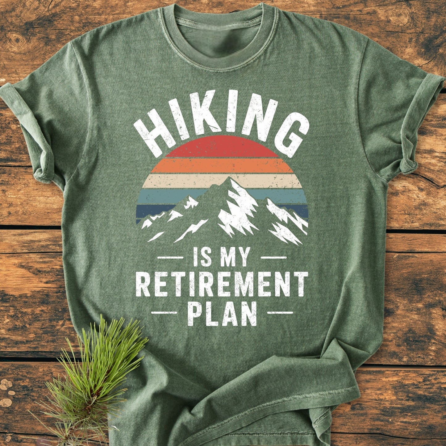 Hiking Is My Retirement Plan - Vintage Wash Tee features a stylized mountain and retro sun. Laid on wood with a pine sprig, showcasing its relaxed fit and durable, soft-washed fabric.