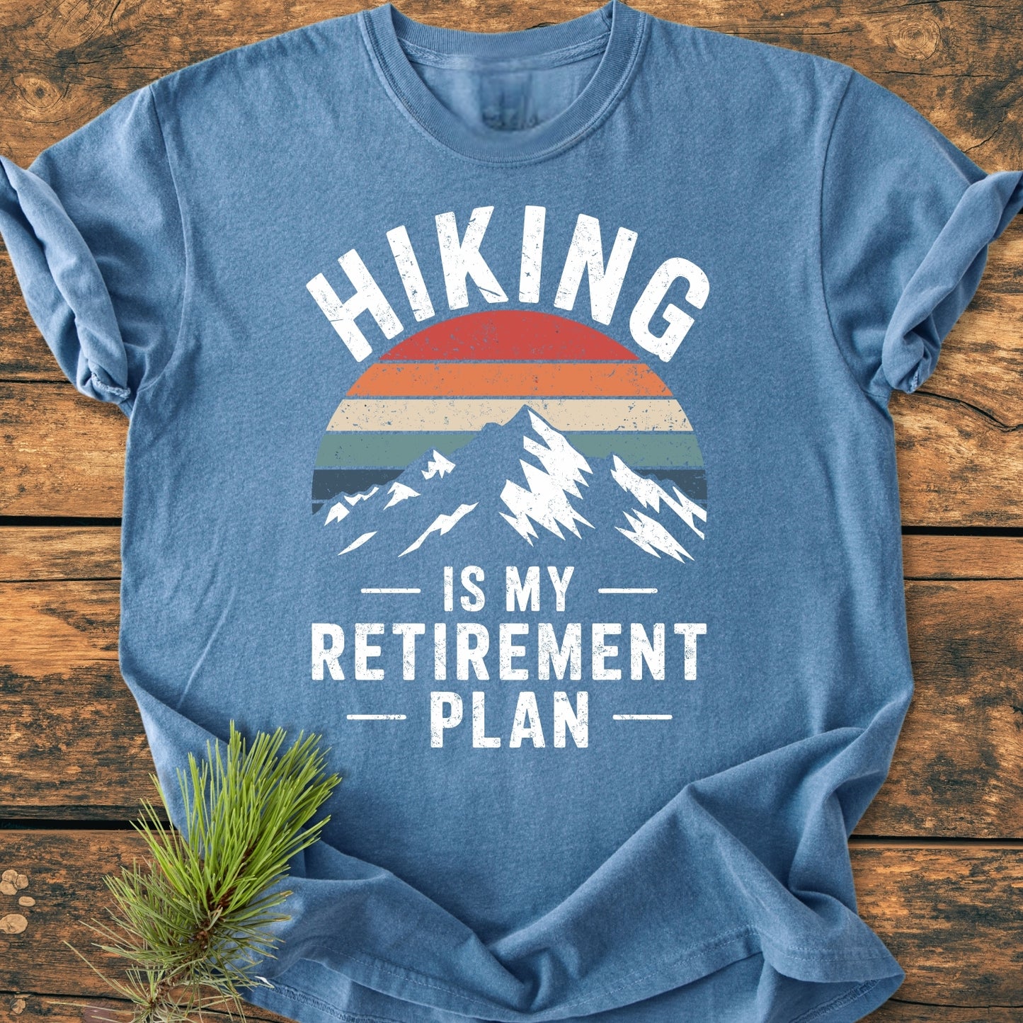 Heather-blue Hiking Is My Retirement Plan - Vintage Wash Tee with a mountain graphic and sunset stripes, laid flat on wood; features bold white text. Pine needles enhance the outdoorsy feel.