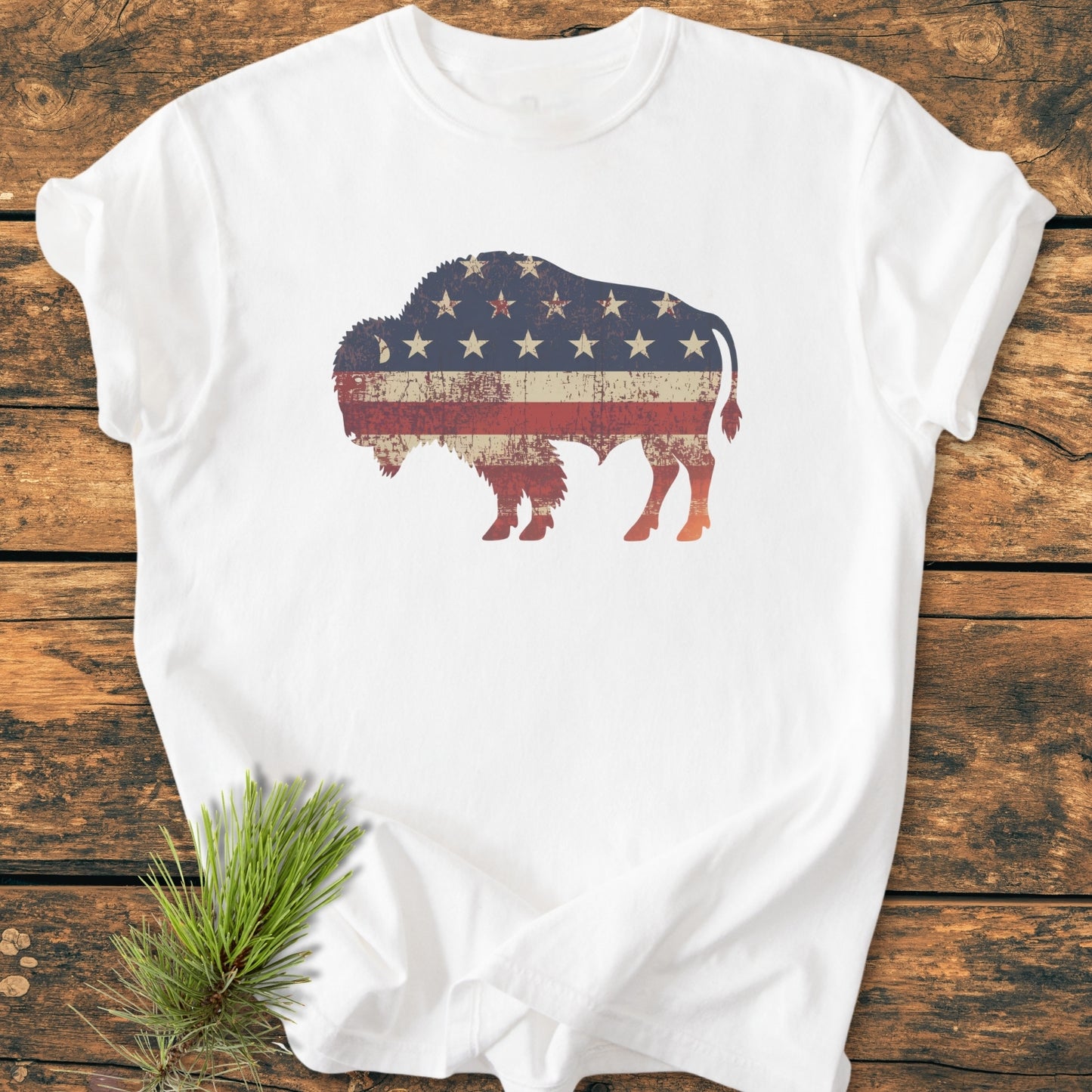 Land Of The Free Bison - Vintage Wash Tee: A white T-shirt featuring a distressed bison silhouette with a vintage U.S. flag motif, laid on rustic wood, accented by a pine sprig.
