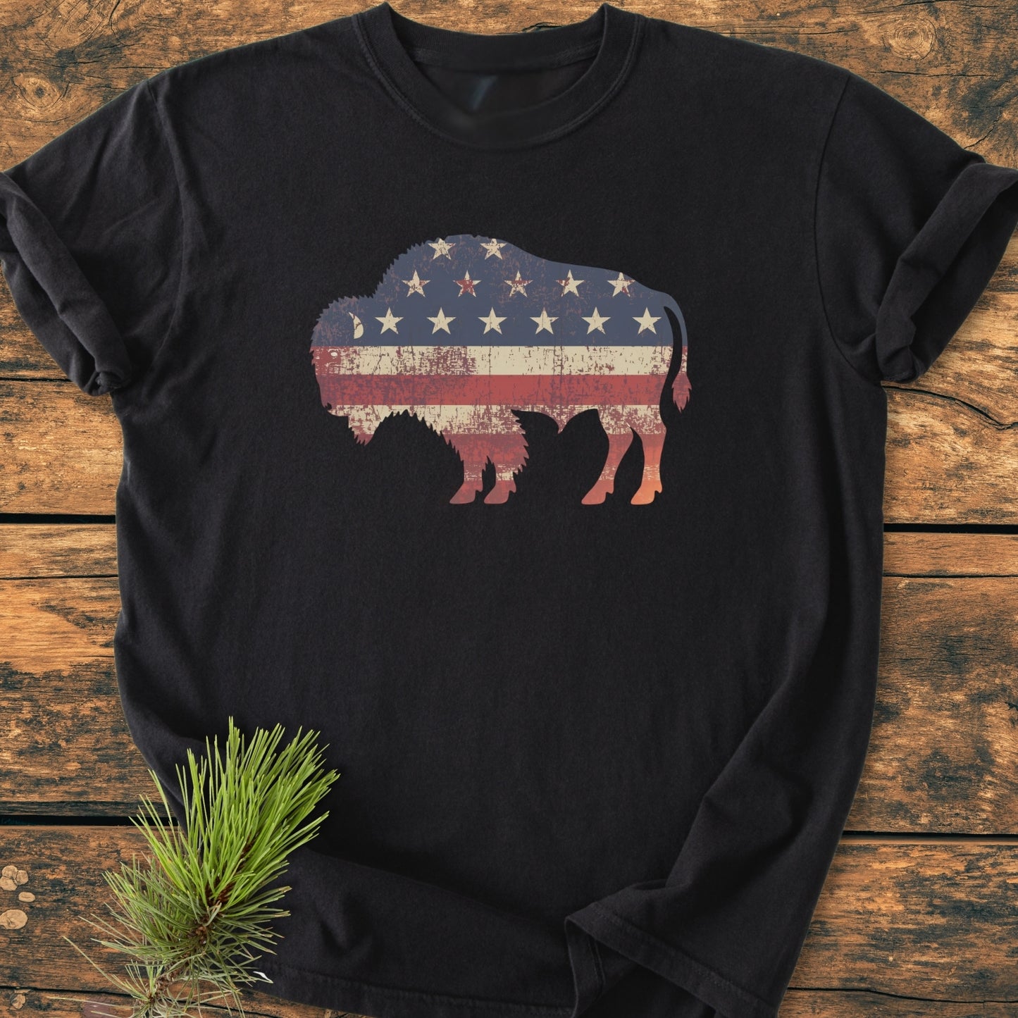 Land Of The Free Bison - Vintage Wash Tee features a buffalo silhouette with a weathered American flag design, laid on a wooden surface with a pine branch accent, showcasing its relaxed fit.