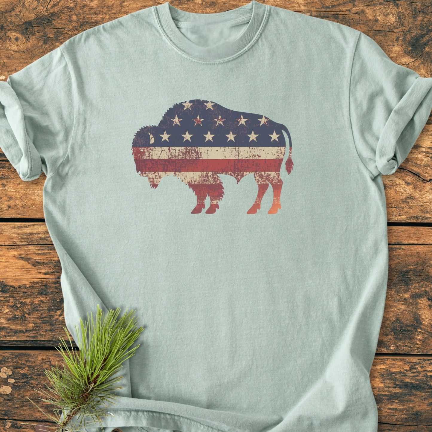 Land Of The Free Bison - Vintage Wash Tee features a distressed bison with a faded U.S. flag motif on a pale-blue Comfort Colors T-shirt, laid on a wooden surface with a small pine sprig.