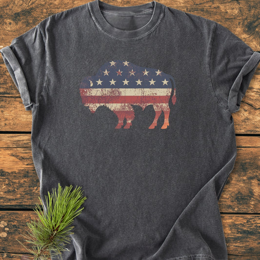 Land Of The Free Bison - Vintage Wash Tee featuring a distressed bison with a U.S. flag motif, laid flat on a wooden surface, styled with a pine needle sprig for a rustic touch.