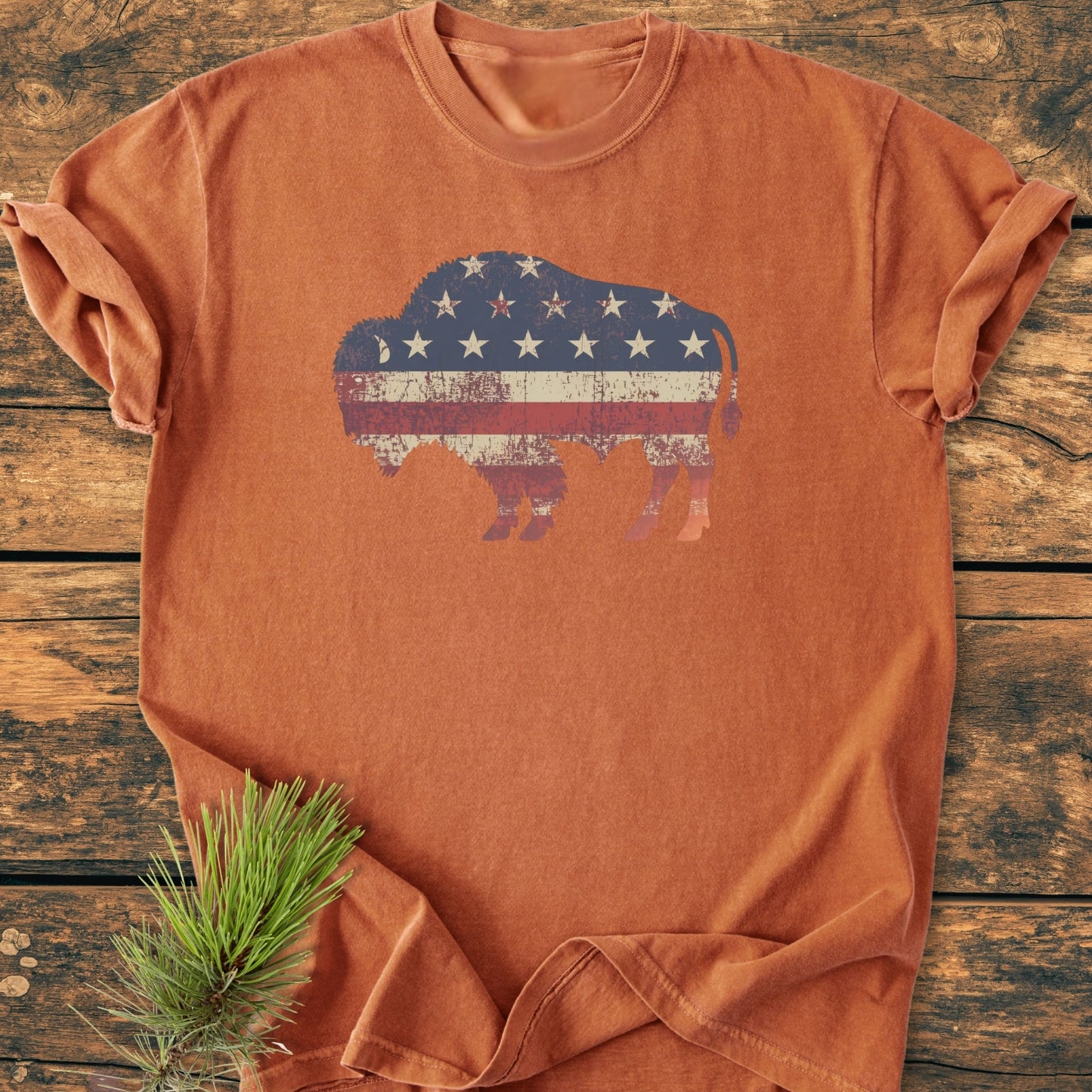 Land Of The Free Bison - Vintage Wash Tee: Earth-tone T-shirt with a distressed bison silhouette featuring an American-flag motif, laid on weathered wood, accompanied by a small pine branch.