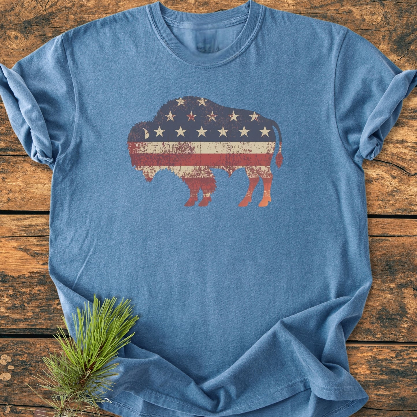 Land Of The Free Bison - Vintage Wash Tee featuring a distressed bison silhouette with an American flag motif on a heathered-blue crew-neck, laid flat on a rustic wooden surface with pine bough accent.