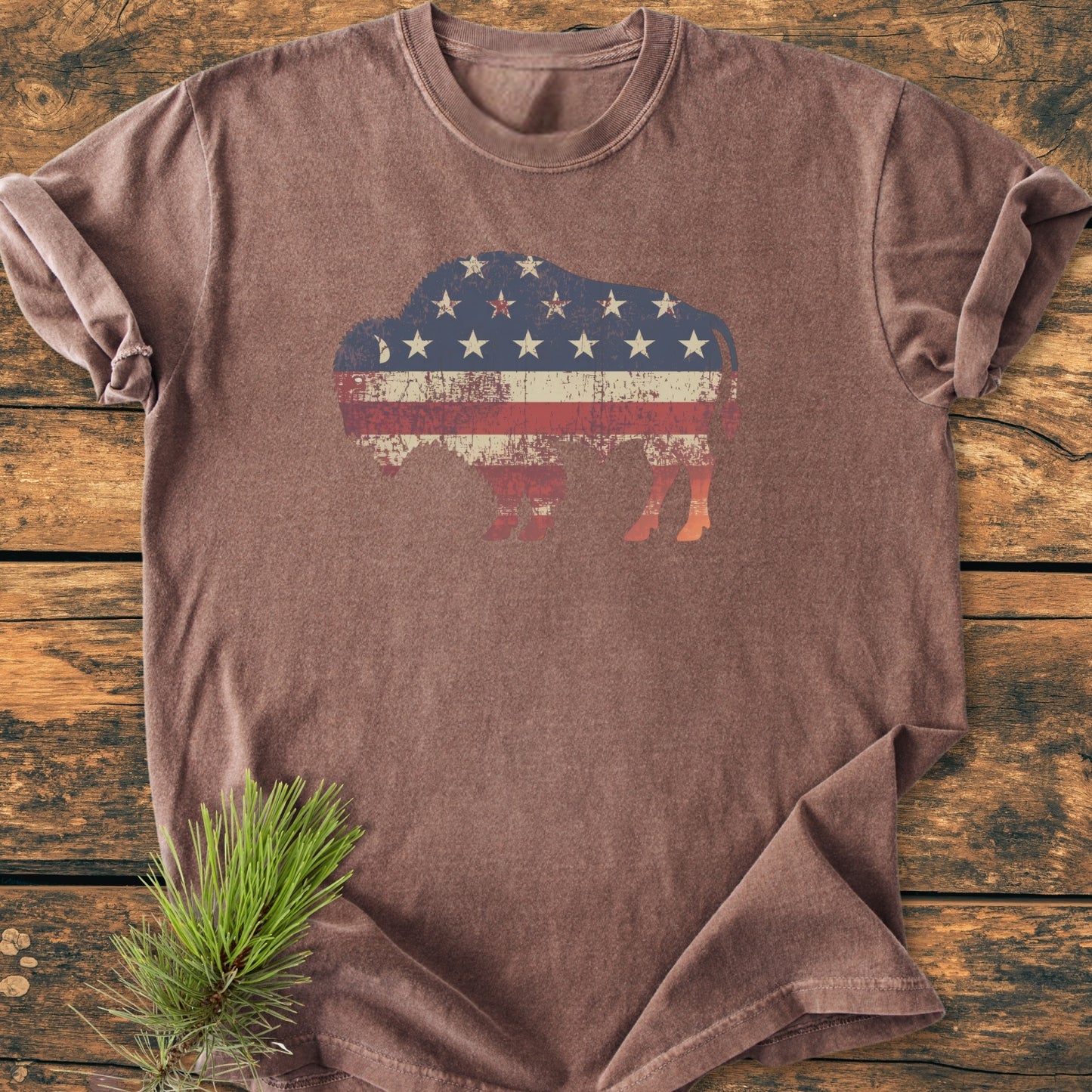 Land Of The Free Bison - Vintage Wash Tee featuring a distressed bison silhouette with U.S. flag design on a brown, short-sleeved shirt, laid on wood with a pine sprig beside it.