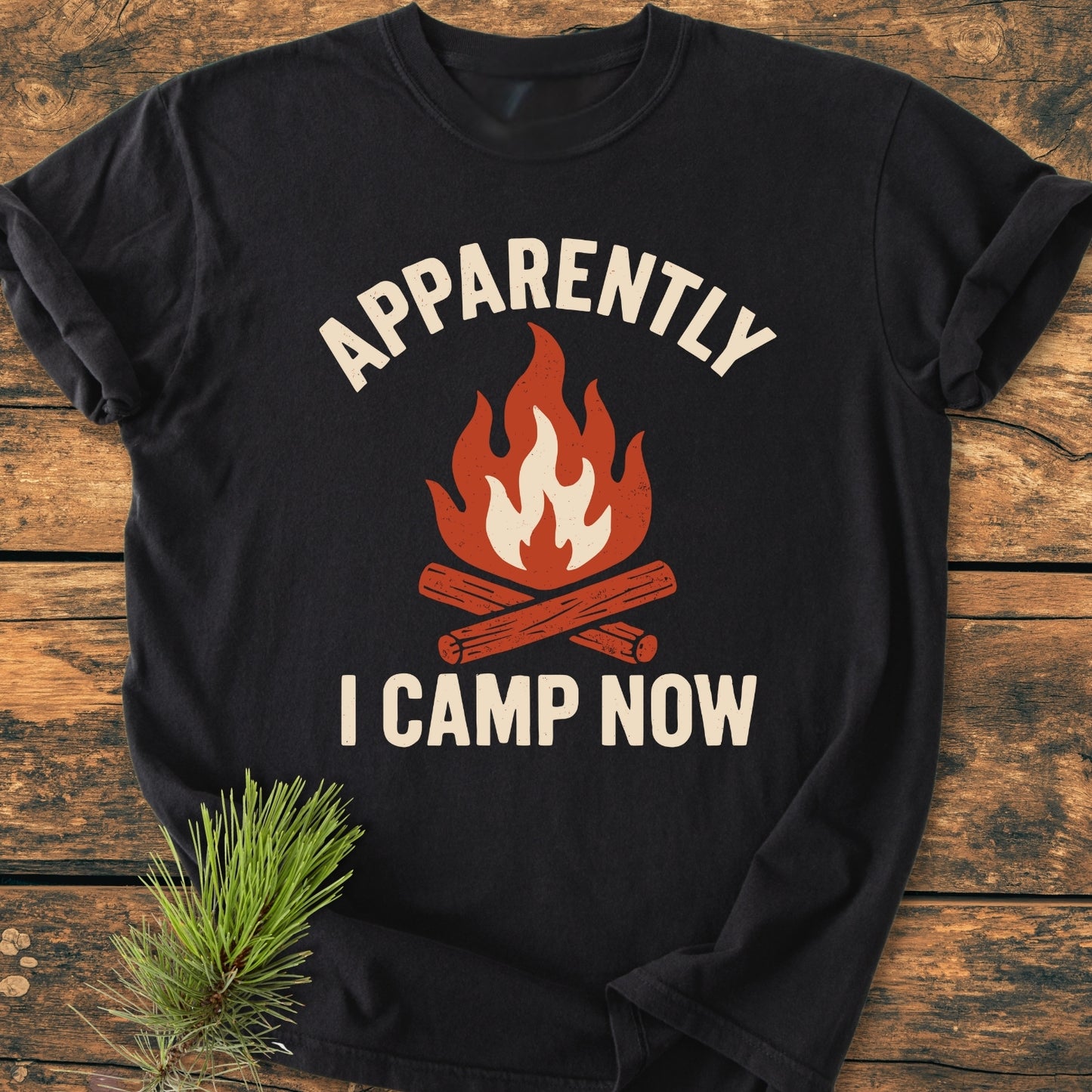 Apparently Camping - Vintage Wash Tee featuring a campfire graphic and slogan, laid on a wooden surface with a pine sprig, highlighting its relaxed fit and soft-washed, garment-dyed fabric.