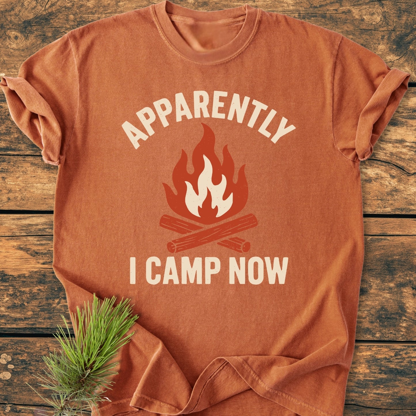Apparently Camping - Vintage Wash Tee: Orange-rust T-shirt with a campfire icon and APPARENTLY and I CAMP NOW lettering. Displayed on wood with pine sprig. Cozy, relaxed fit, garment-dyed cotton.