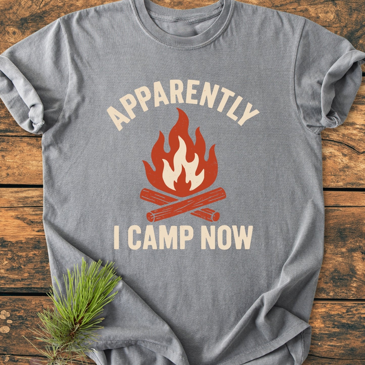 Apparently Camping - Vintage Wash Tee featuring a campfire graphic with crossed logs and flames, laid on rustic wood, accompanied by a pine sprig, showcasing its cozy, relaxed fit and durable design.