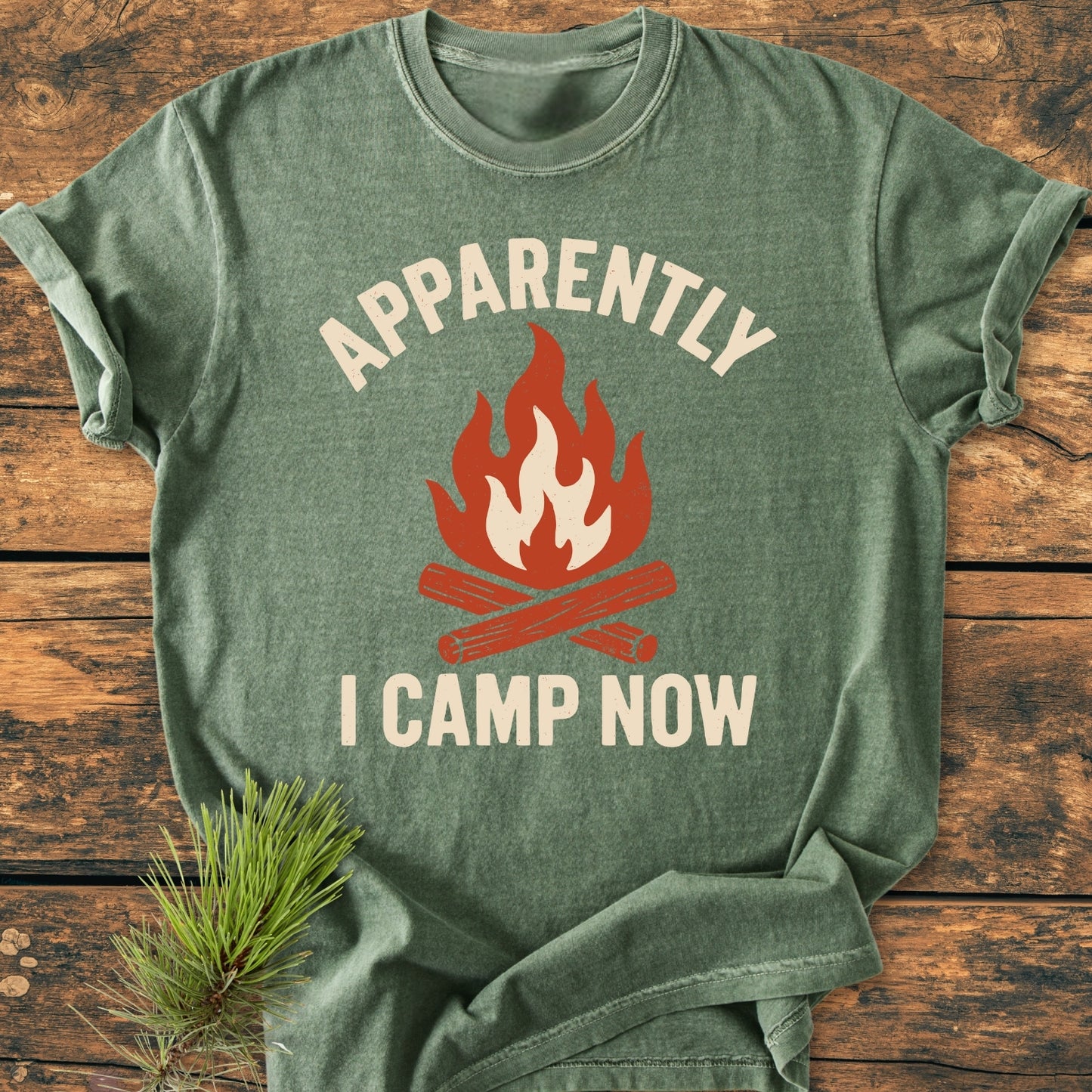 Apparently Camping - Vintage Wash Tee features a heather-green color with a stylized campfire graphic and humorous text, APPARENTLY I CAMP NOW, on a rustic wooden background with an evergreen sprig.