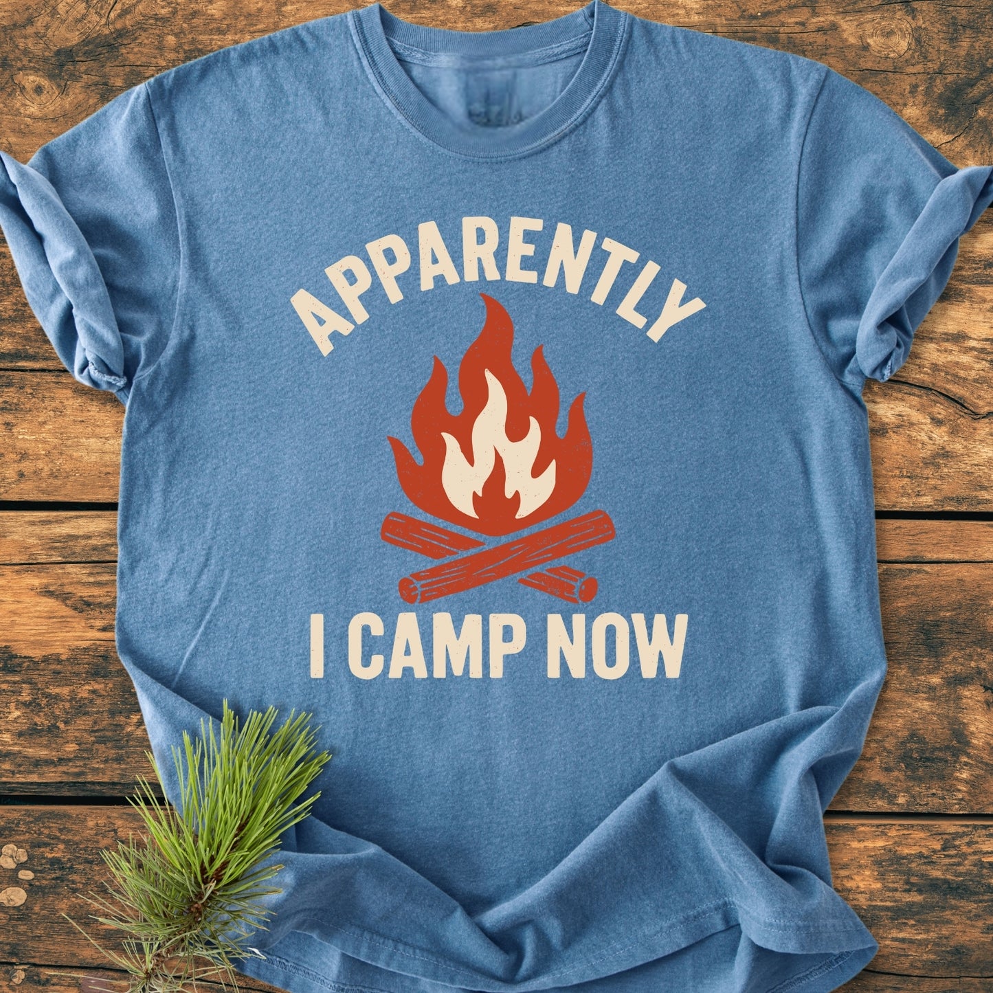Apparently Camping - Vintage Wash Tee features a campfire graphic and bold text on a blue T-shirt, laid flat with a pine branch accent, highlighting its relaxed fit and soft-washed cotton fabric.