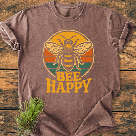 Bee Happy - Vintage Wash Tee features a retro bee graphic with BEE HAPPY slogan on a brown, soft-washed, garment-dyed T-shirt, laid out on a wooden surface with a pine sprig beside it.