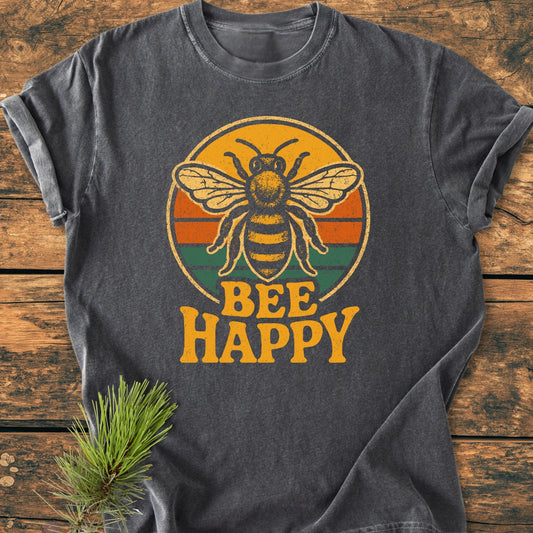 Bee Happy - Vintage Wash Tee features a charcoal-gray design with a retro bee graphic and slogan “BEE HAPPY” on soft, garment-dyed cotton, styled on a rustic wood surface with a pine sprig.