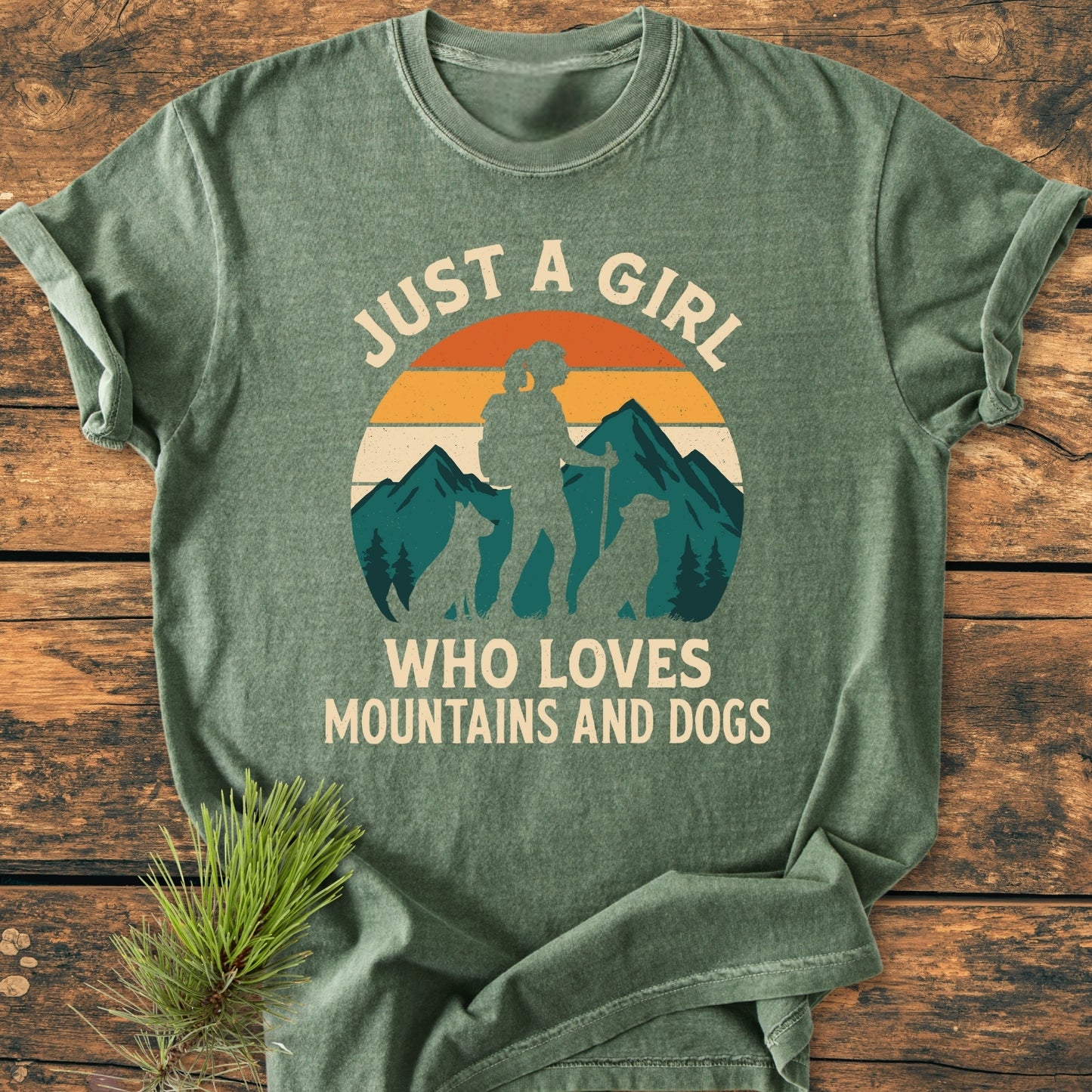 Mountains And Dogs - Vintage Wash Tee: Heathered-green T-shirt featuring a retro mountain graphic with hiker and dogs, surrounded by the phrase Just a Girl Who Loves Mountains and Dogs, laid on wood.