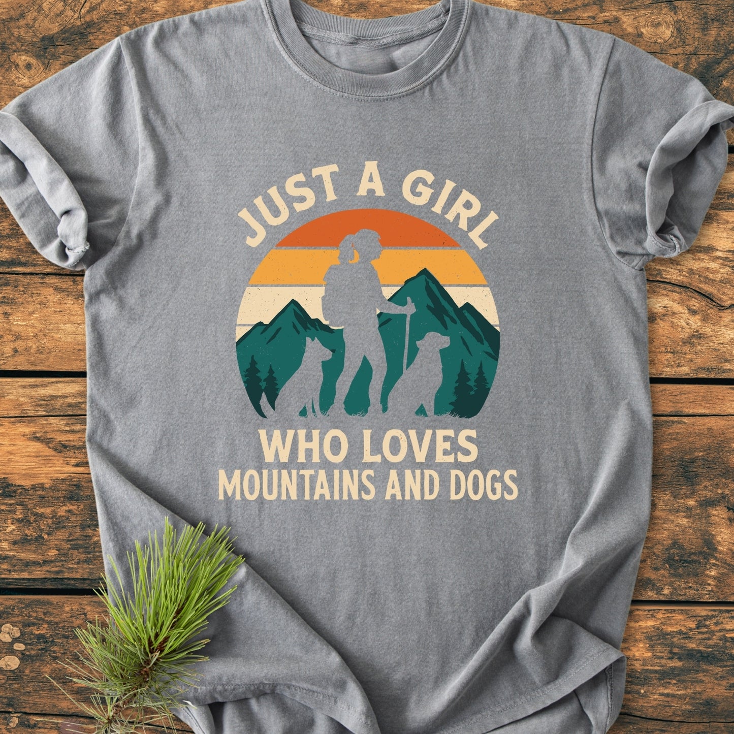 Mountains And Dogs - Vintage Wash Tee features a circular graphic of a girl hiking with dogs against a mountain sunset, laid on wood with a pine sprig, showcasing its relaxed fit and durability.