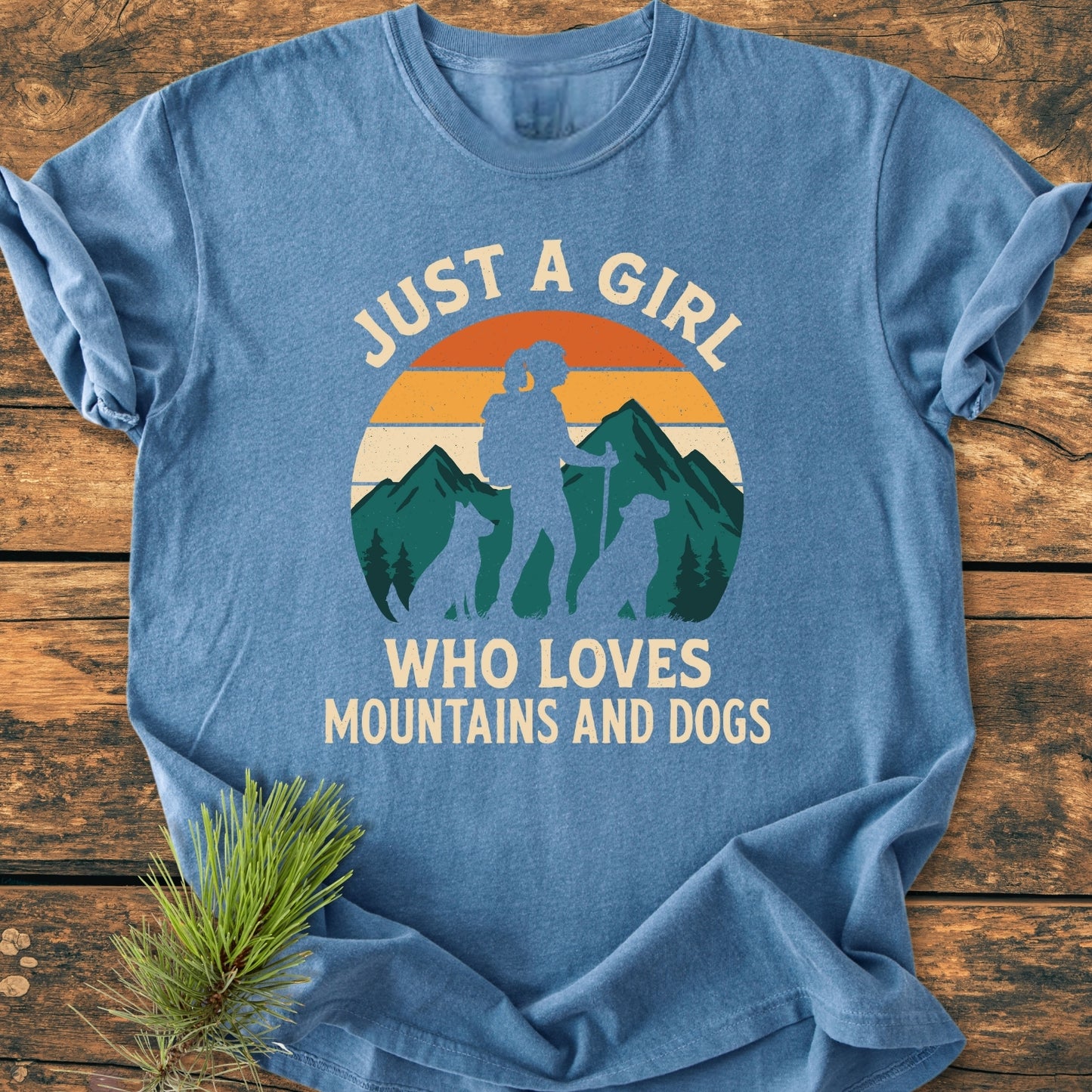 Mountains And Dogs - Vintage Wash Tee featuring a retro graphic of a girl with dogs, mountains, and sunset, laid on wood. Soft, relaxed fit, durable with double-needle stitching, 100% ring-spun cotton.