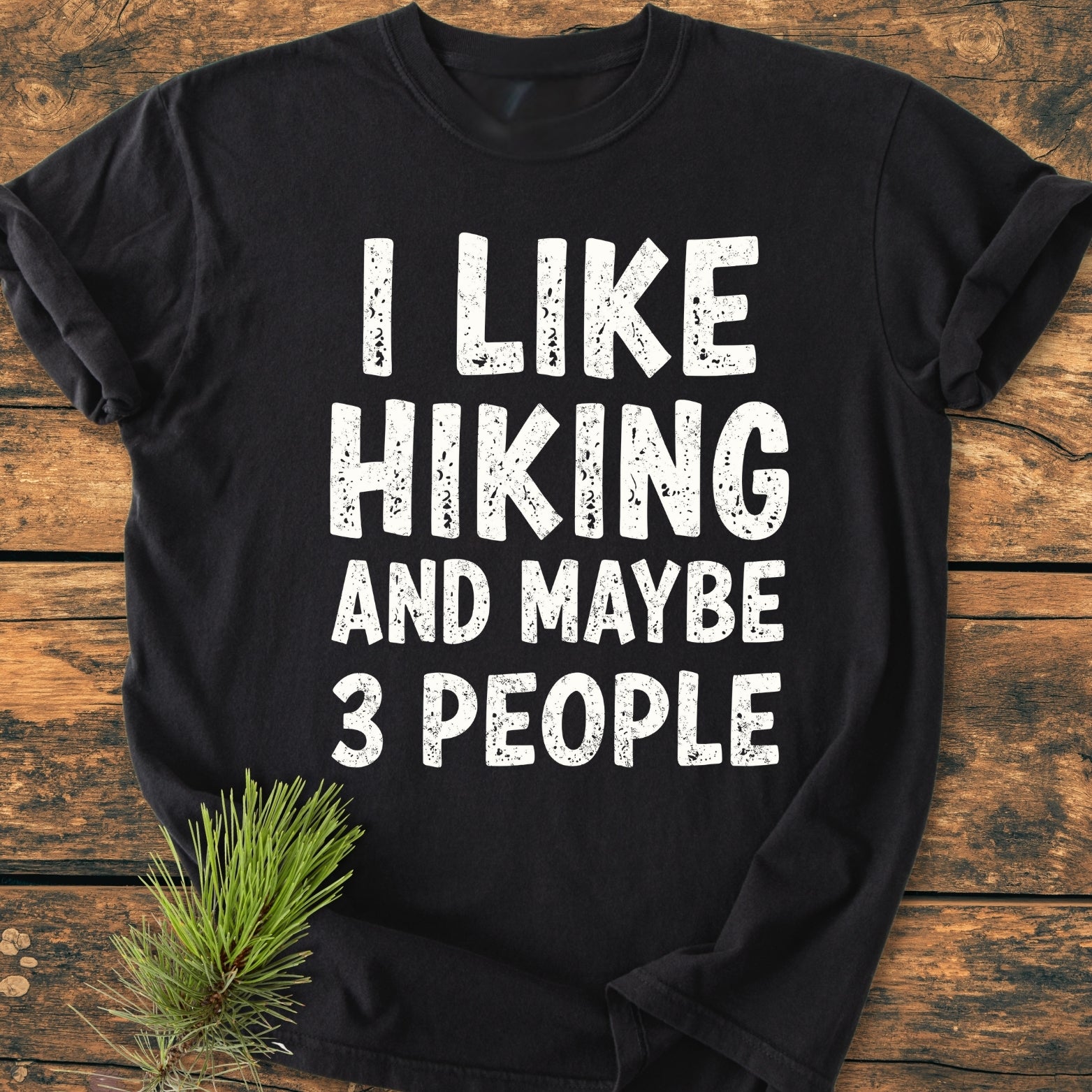 I Like Hiking And Maybe 3 People - Vintage Wash Tee, black, laid flat on wood with a pine sprig, features a humorous slogan in large distressed white print, embodying comfort and style.