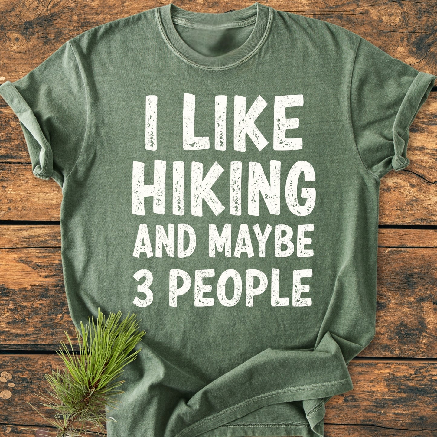 Heather-green T-shirt with I LIKE HIKING AND MAYBE 3 PEOPLE slogan in distressed white letters, laid flat on wood. Features soft garment-dyed fabric, providing a relaxed fit. Pine sprig beside it.