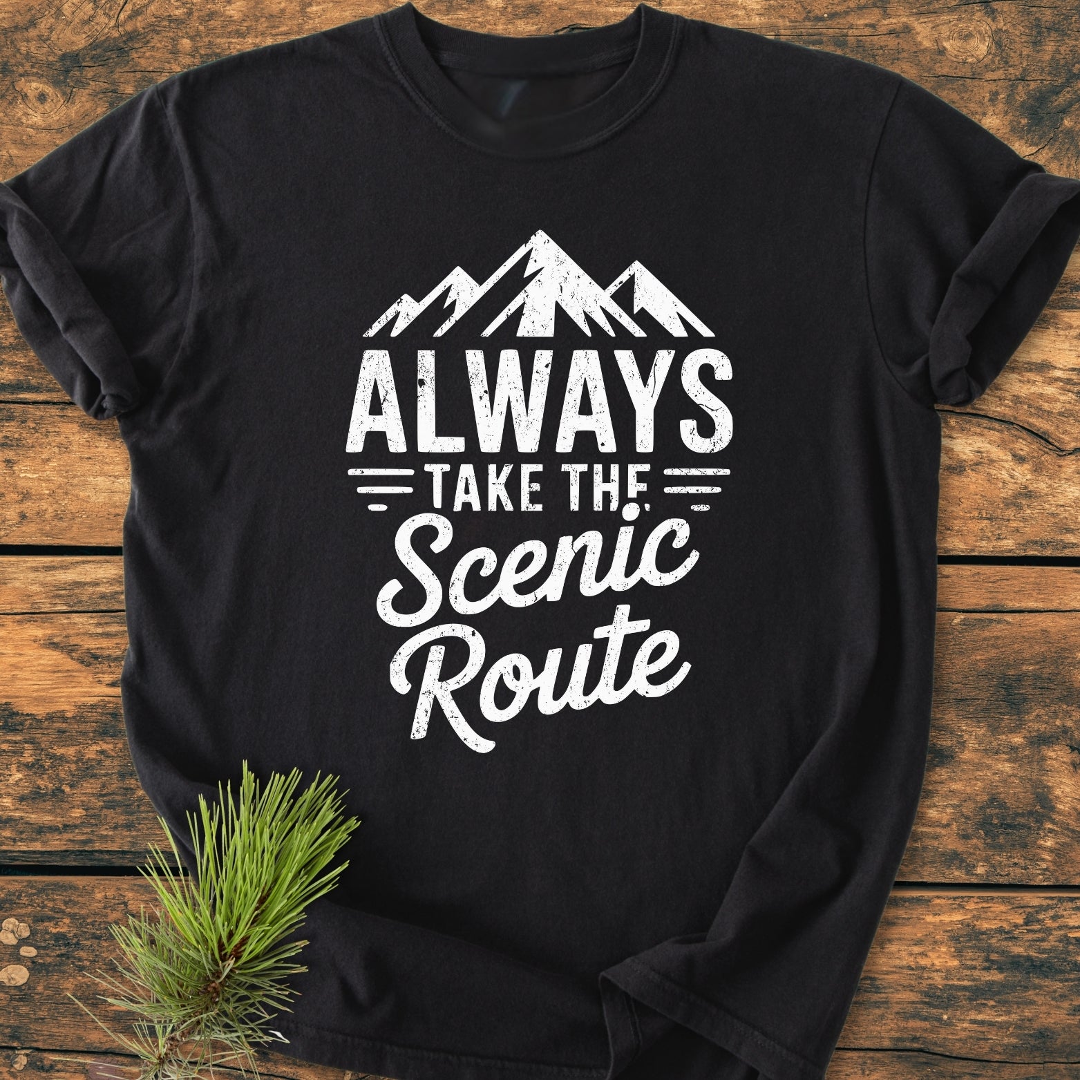 Always Take The Scenic Route - Vintage Wash Tee: Black short-sleeve T-shirt with white distressed lettering and mountain graphic, laid flat on wooden surface, accented by a pine branch. Relaxed fit, durable, garment-dyed cotton.