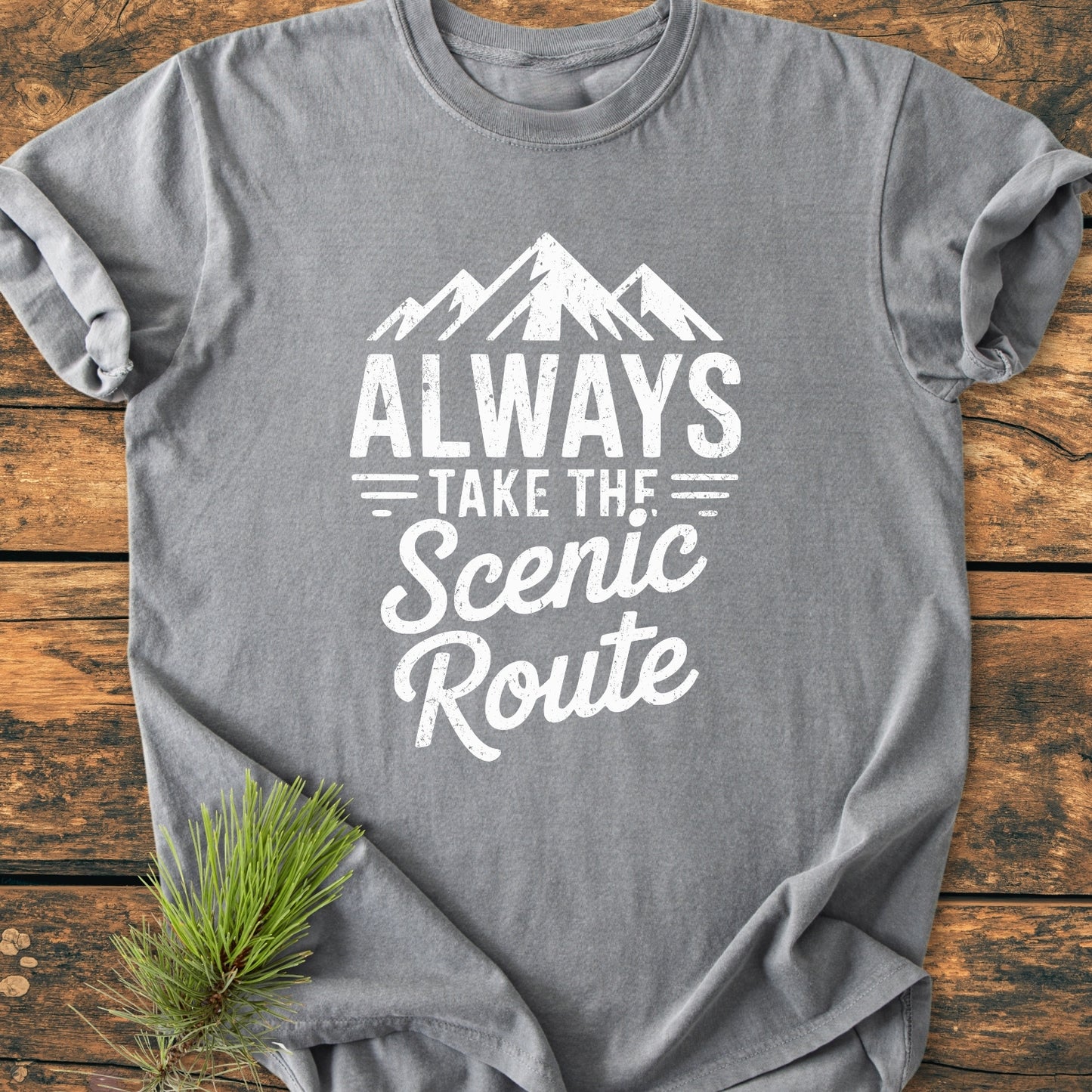 Always Take The Scenic Route - Vintage Wash Tee features a heather-gray color with a mountain graphic and slogan, laid on wood. Pine needles add outdoor charm, showcasing its relaxed fit and durability.