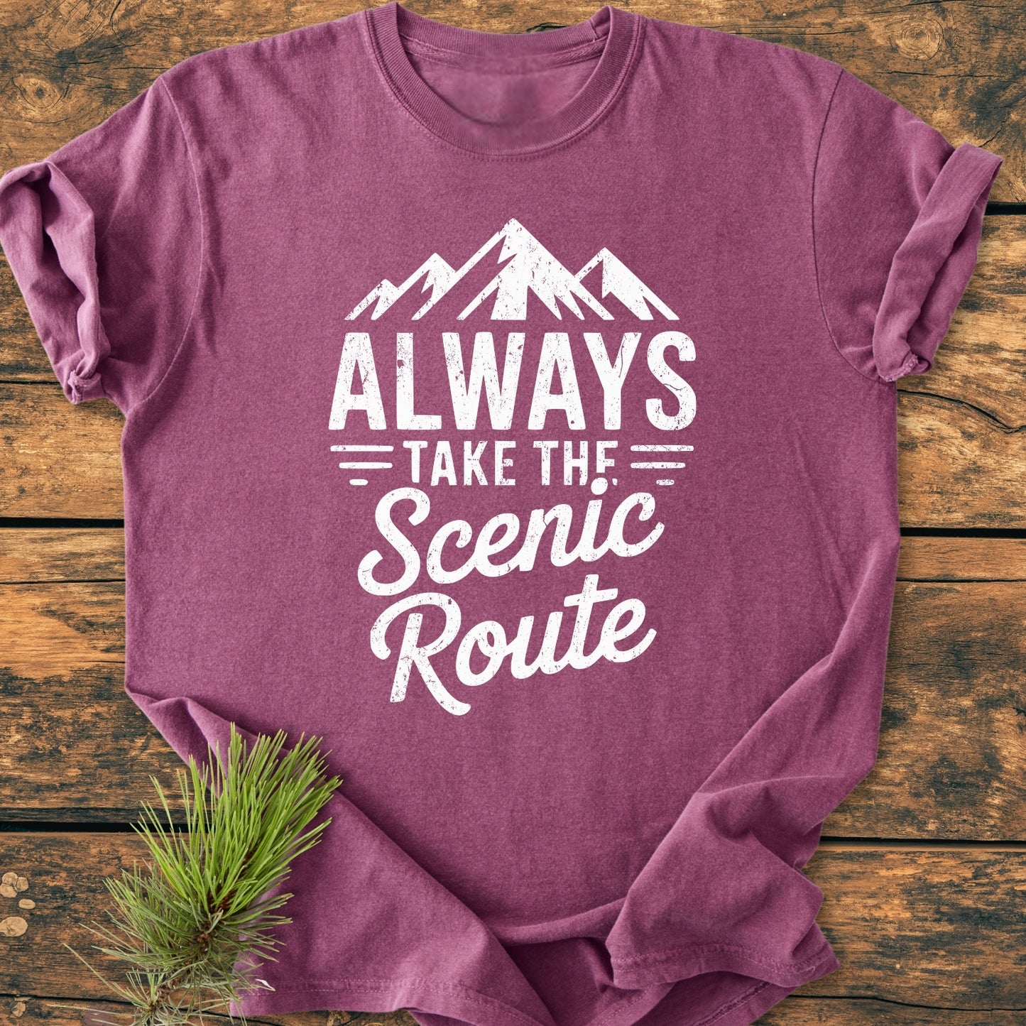 Always Take The Scenic Route - Vintage Wash Tee, featuring white mountain peaks graphic and slogan on a heathered maroon T-shirt, laid flat on a wooden surface with a pine sprig nearby.