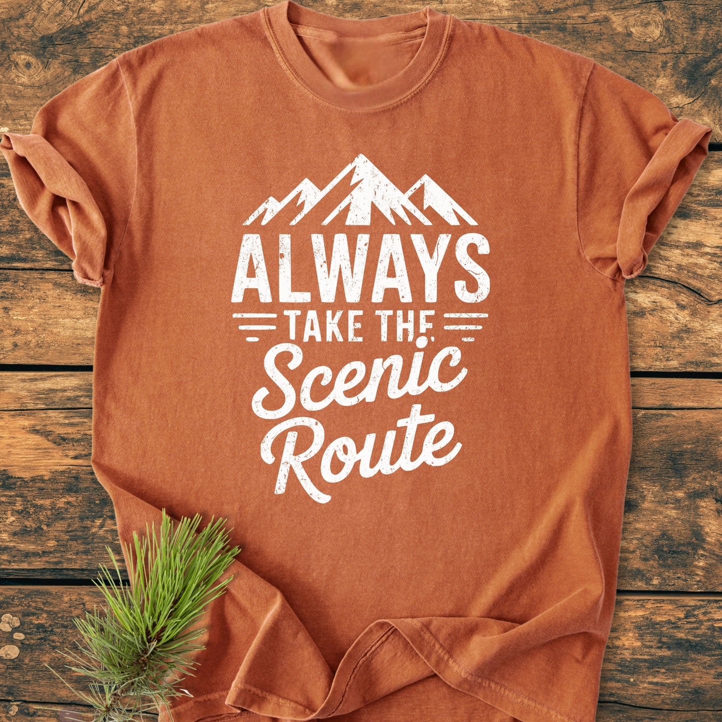 Always Take The Scenic Route - Vintage Wash Tee featuring a mountain silhouette slogan on rust-orange fabric, laid on a wooden surface with a pine sprig, showcasing its relaxed, soft-washed, durable design.