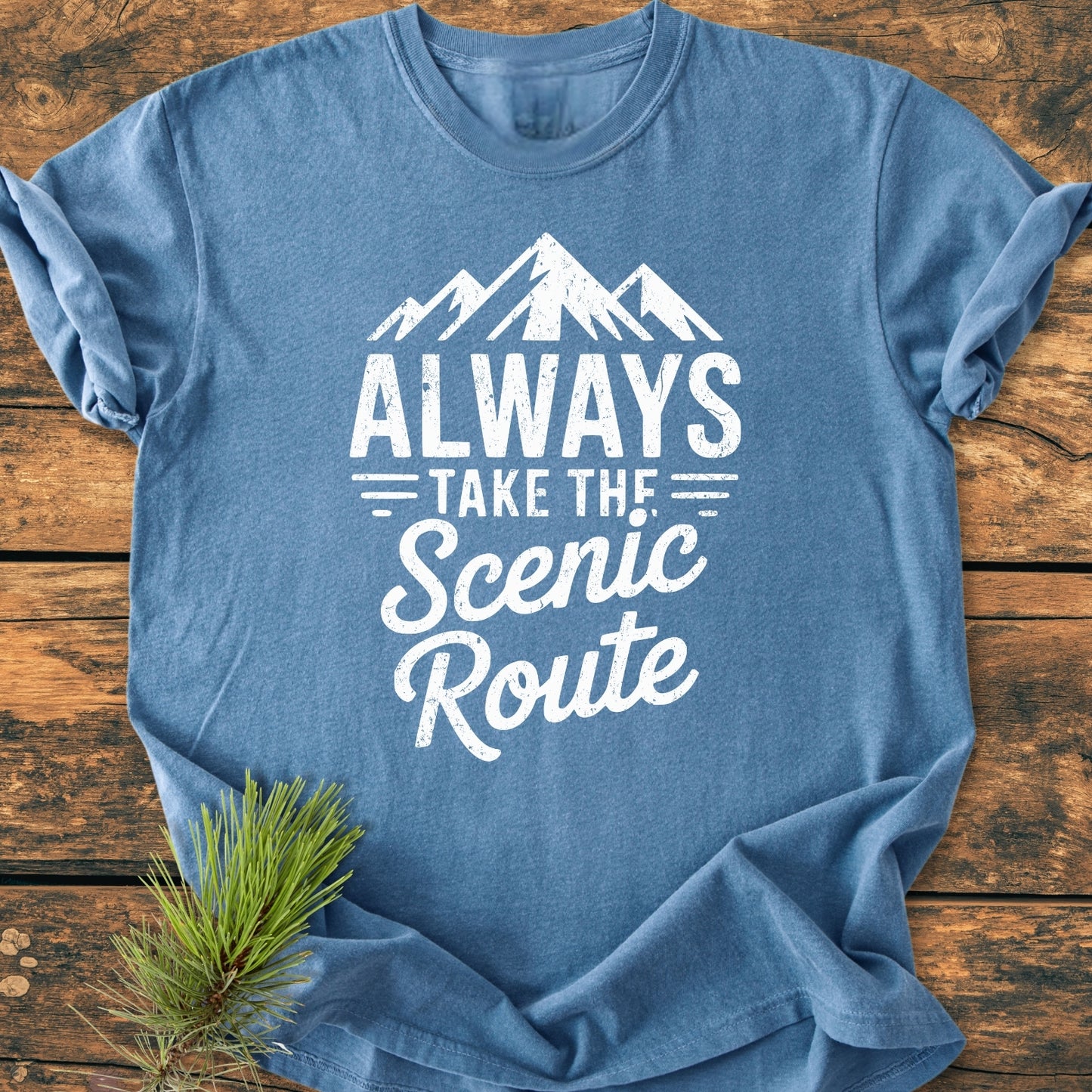 Always Take The Scenic Route - Vintage Wash Tee features a distressed mountain graphic and slogan on a heather-blue shirt, laid on wood with a pine branch accent.
